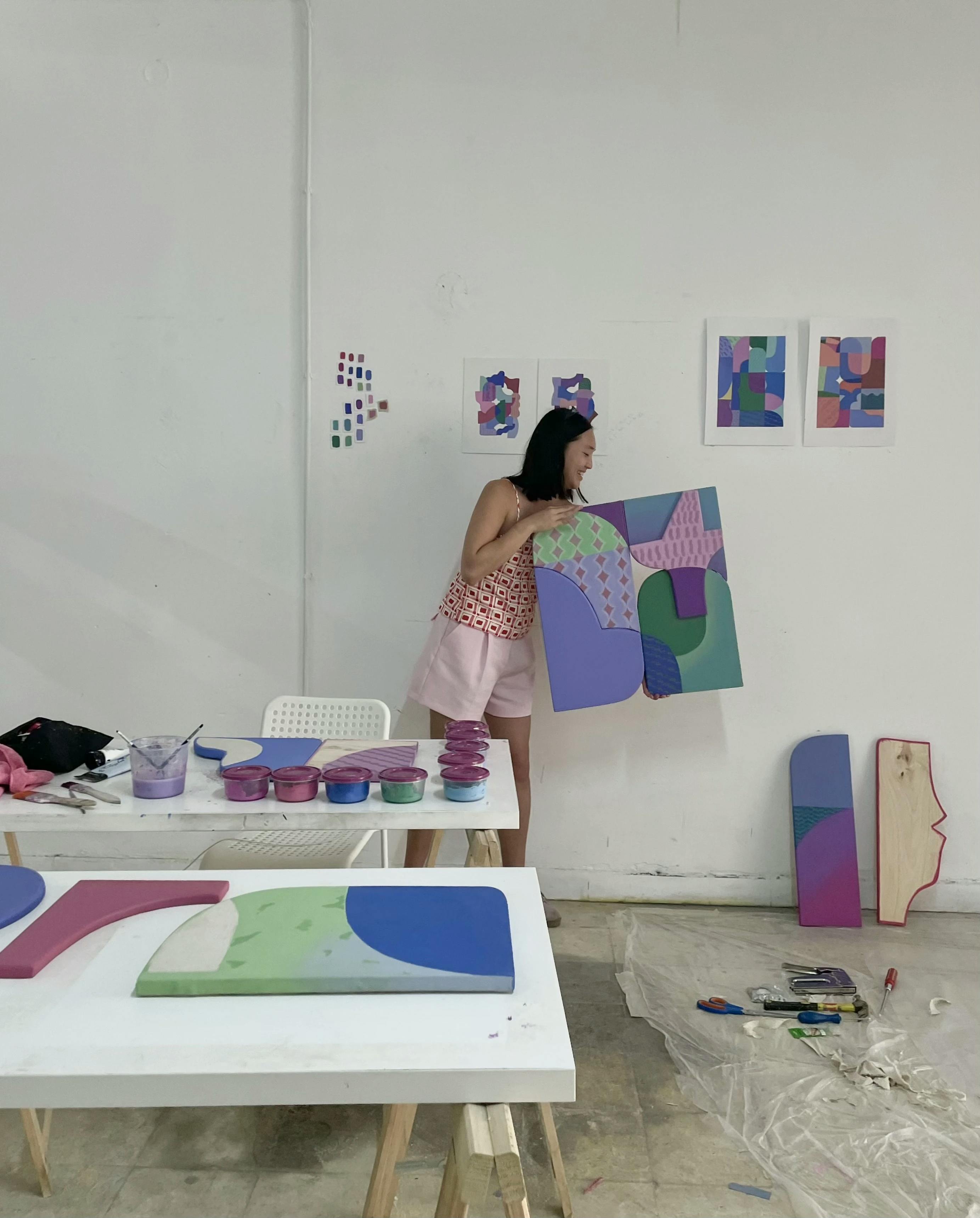 Artist Bbblob holding a colorful shaped canvas with geometric patterns in her studio.