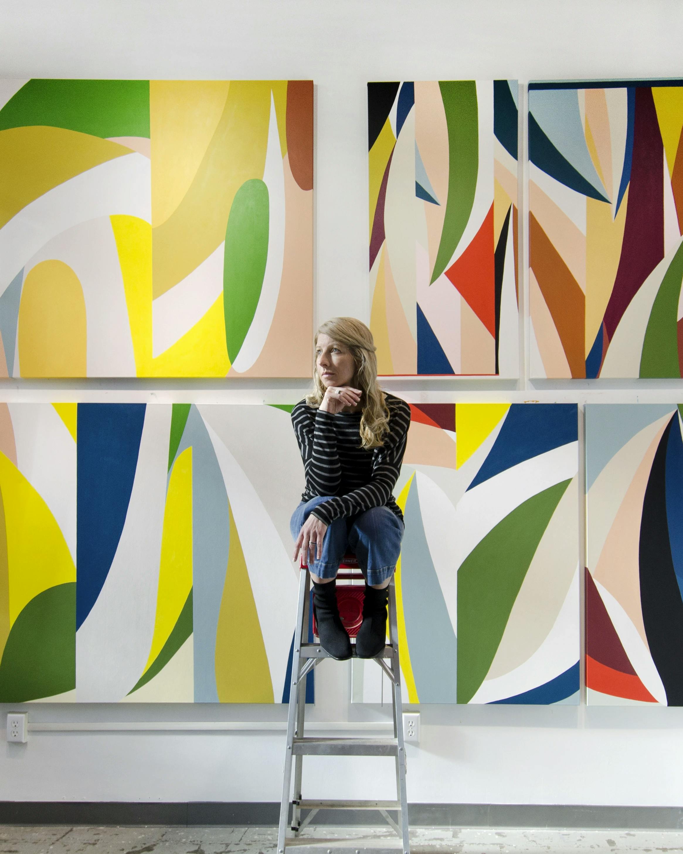 Artist Christina Flowers sitting on a ladder in front of her large colorful paintings. 