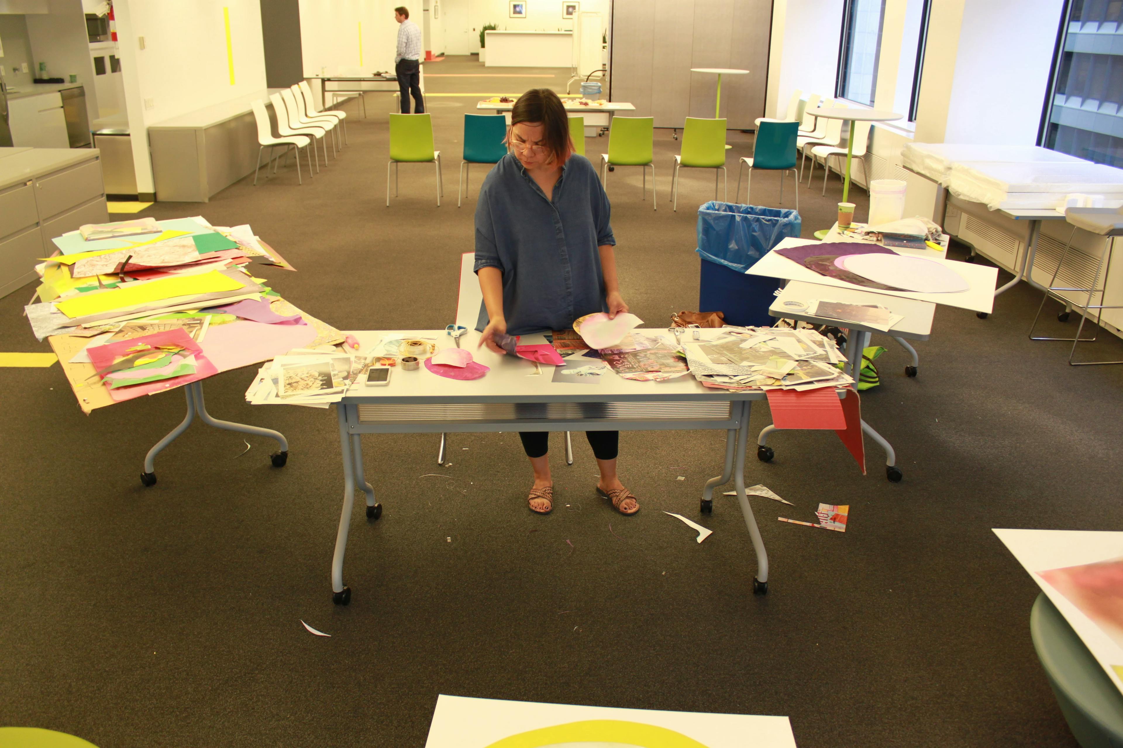Artist Xochi Solis in an office building standing in front of three desks covered with scraps of paper and collage material.