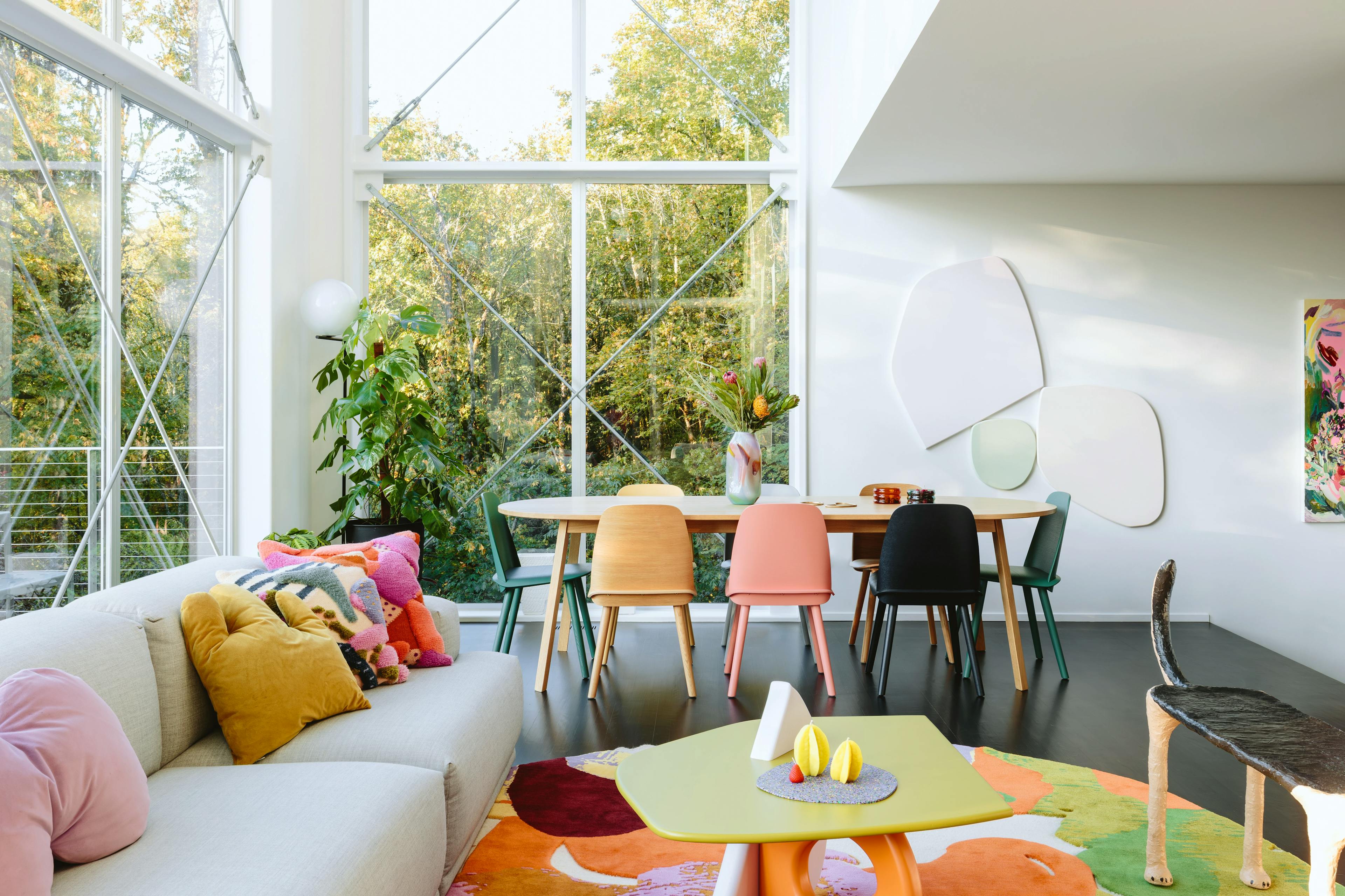 A light-filled sunroom with floor-to-ceiling windows, gray couch, and dining table with colorful, mismatched chairs.