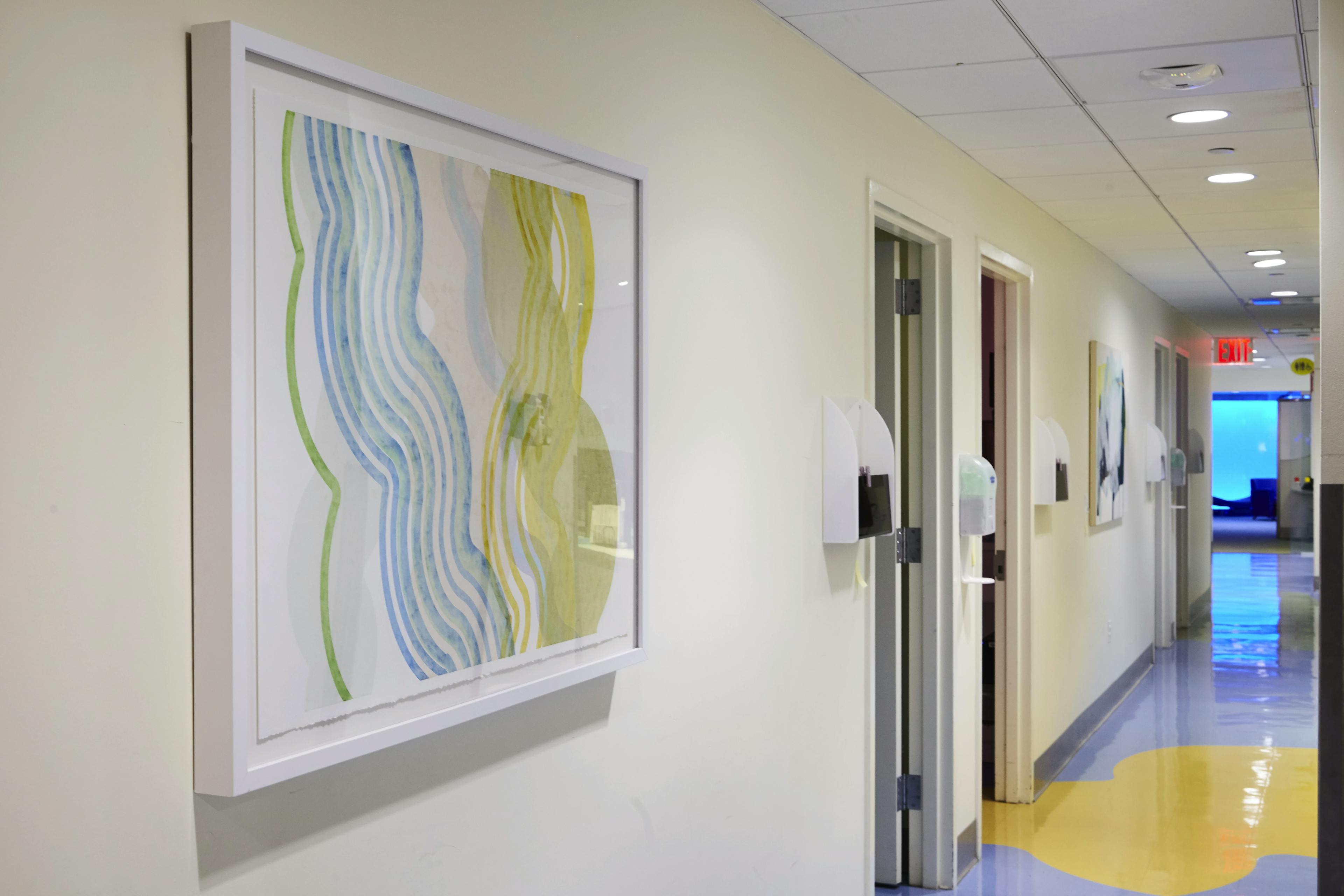 Framed artwork with striated curves installed on the beige wall of a hospital corridor.
