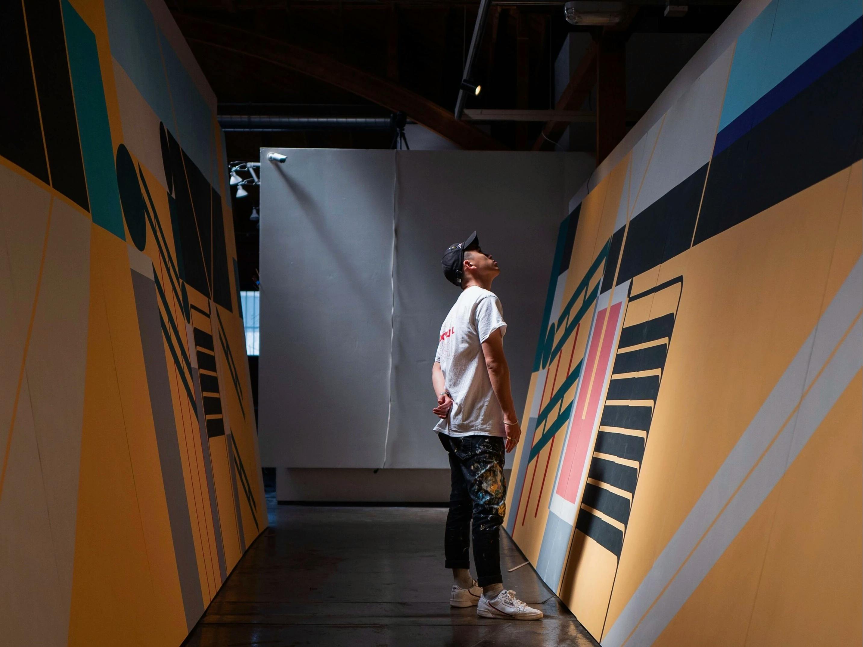 Artist Adrian Kay Wong wearing a baseball cap, white shirt, and paint-splattered jeans, standing in-between his two multicolored wall murals.