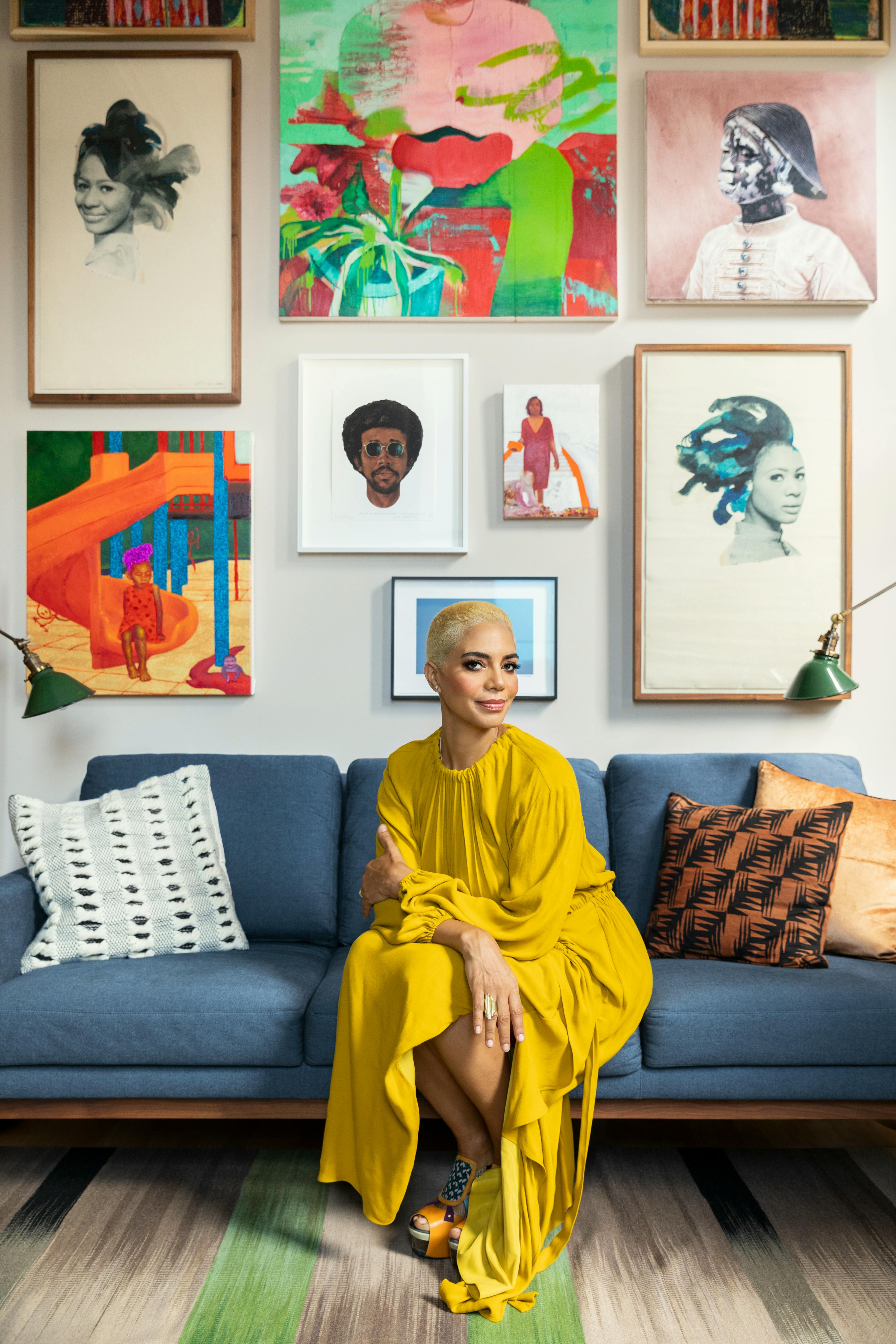 A portrait of interior designer Danielle Colding, wearing a long yellow dress and seated on a blue couch with multiple artworks hanging behind her.
