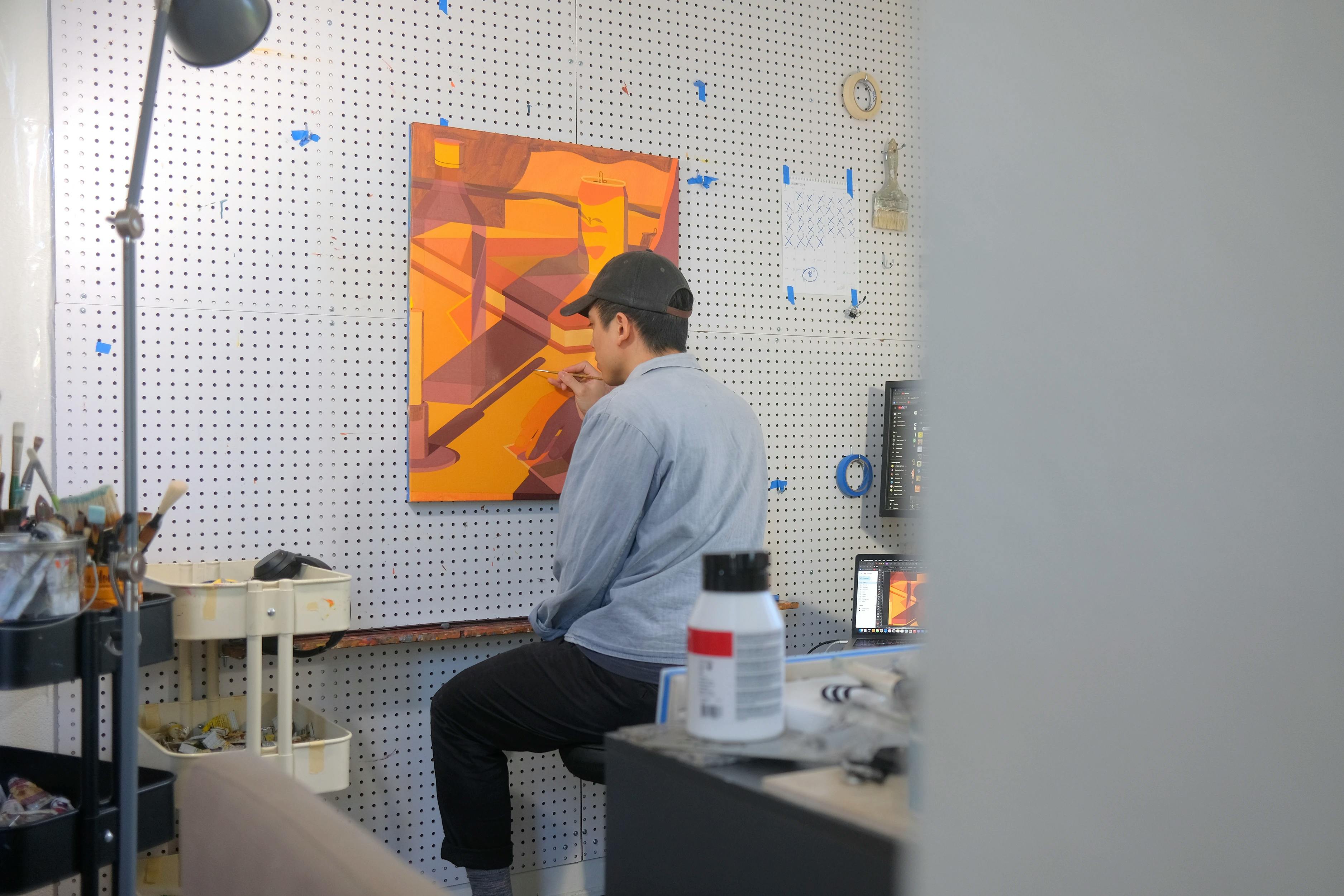Artist Adrian Kay Wong sitting, working on an orange painting in his studio. 
