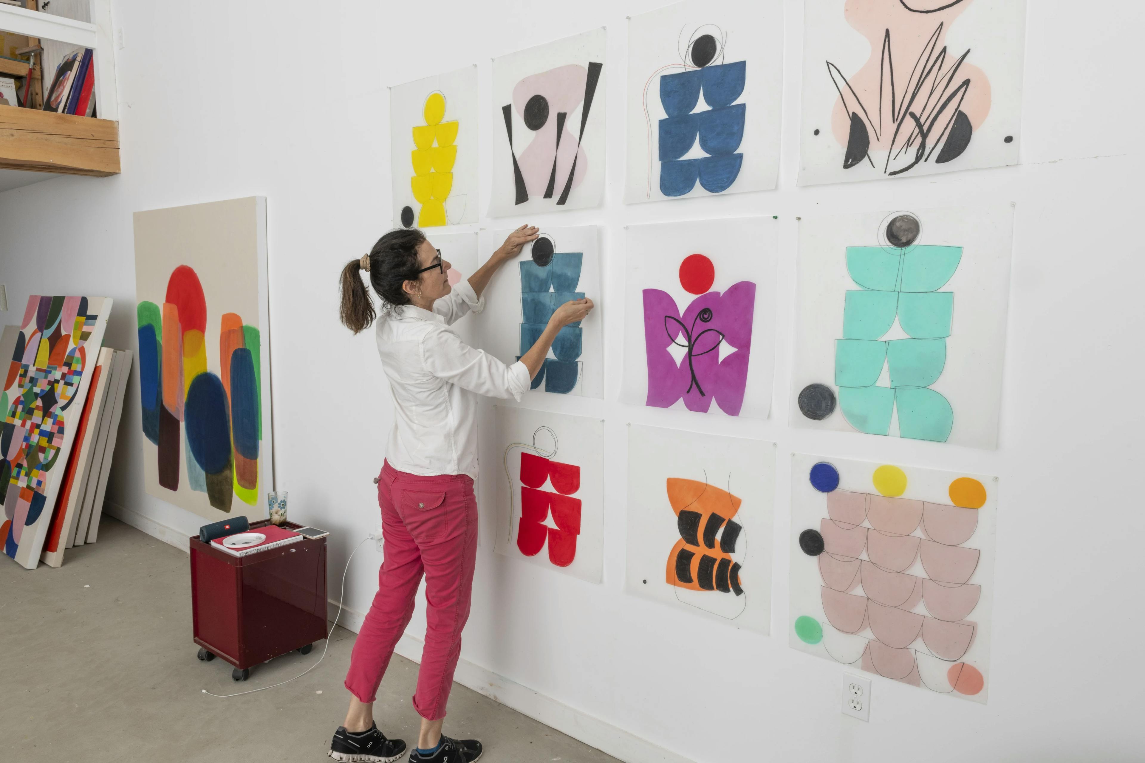 Artist Vicki Sher standing in front of a grid of her abstract, colorful paintings pinned to a white wall in her studio.