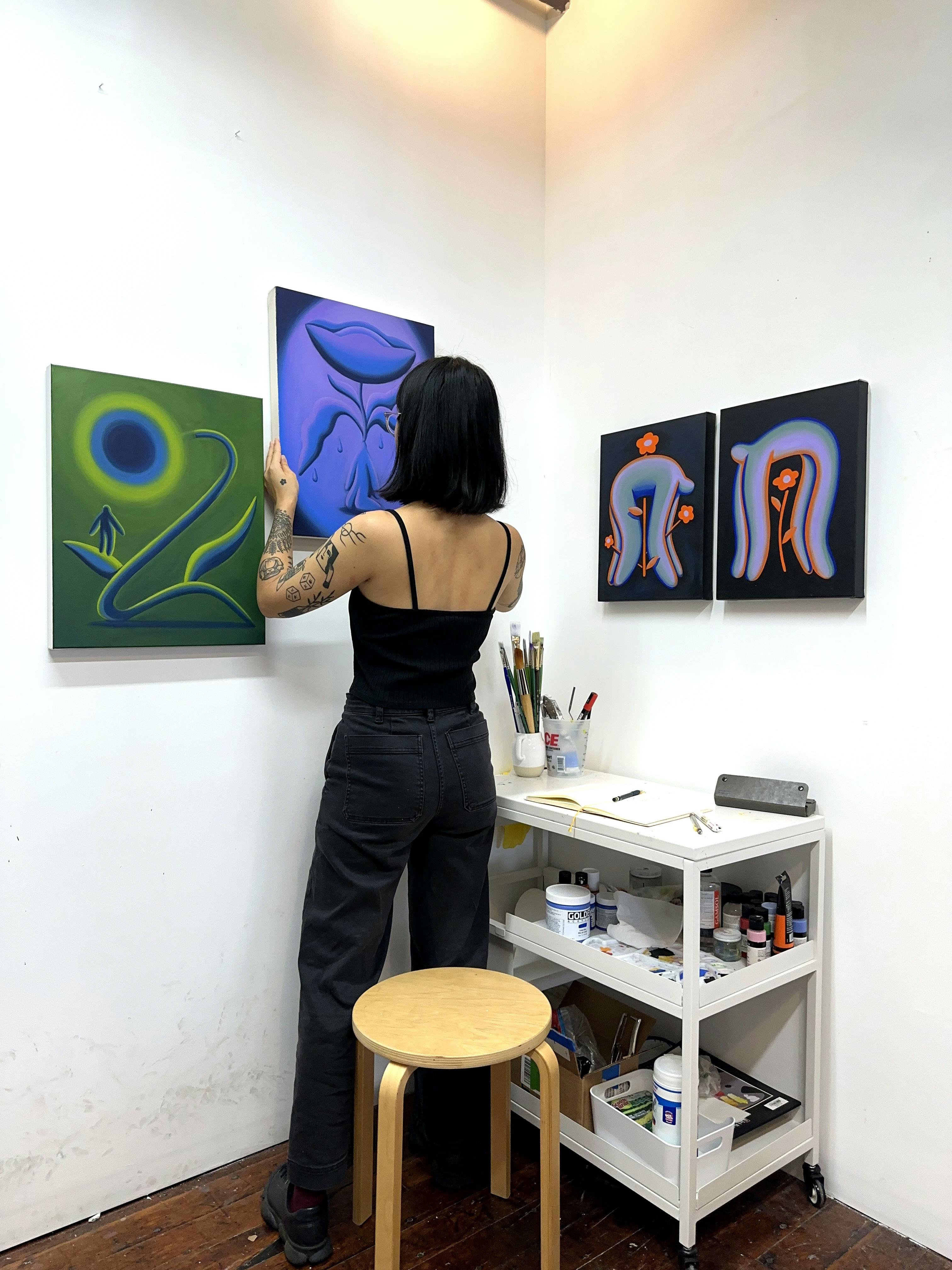 Artist Jocelyn Tsaih hanging up a purple painting next to other paintings on a white wall in her studio.