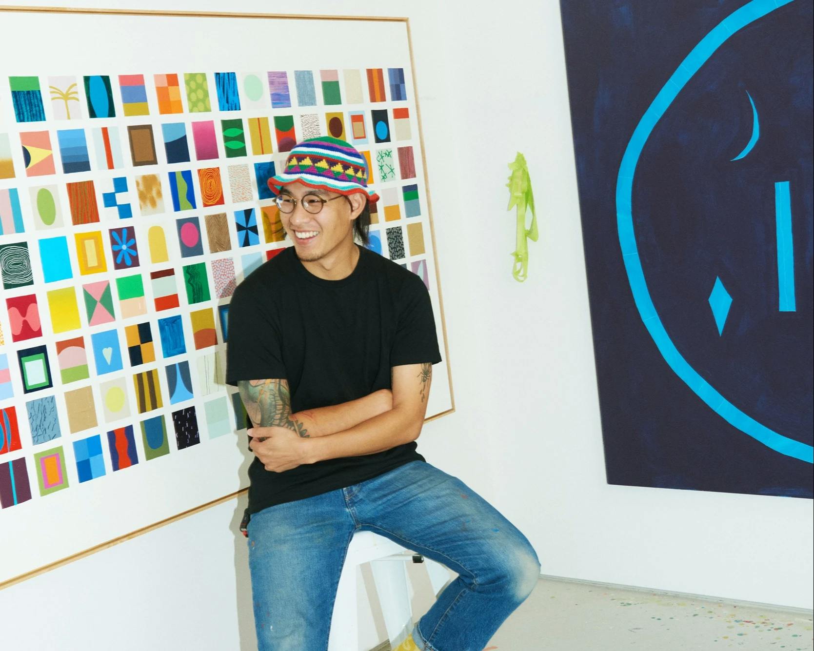 Artist Scott Sueme sitting on a stool in his studio surrounded by large, abstract paintings.