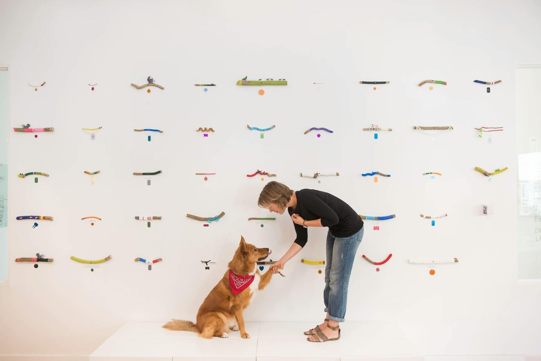 Artist Christina Watka standing in front of a wall with colorful stick sculptures, holding the paw of a dog with a red bandana.