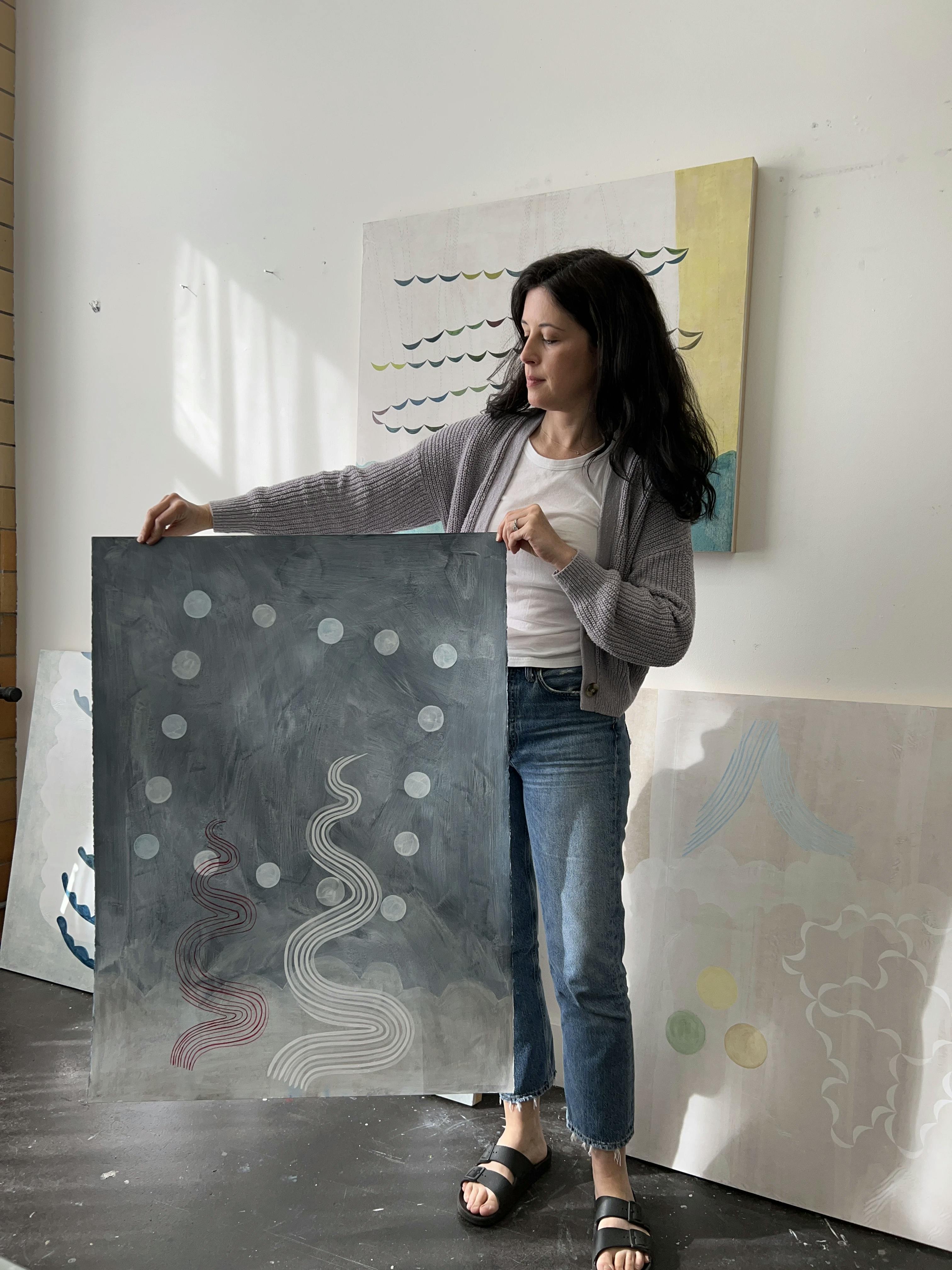 Artist Lydia Bassis holding a light gray abstract painting on paper in her studio.