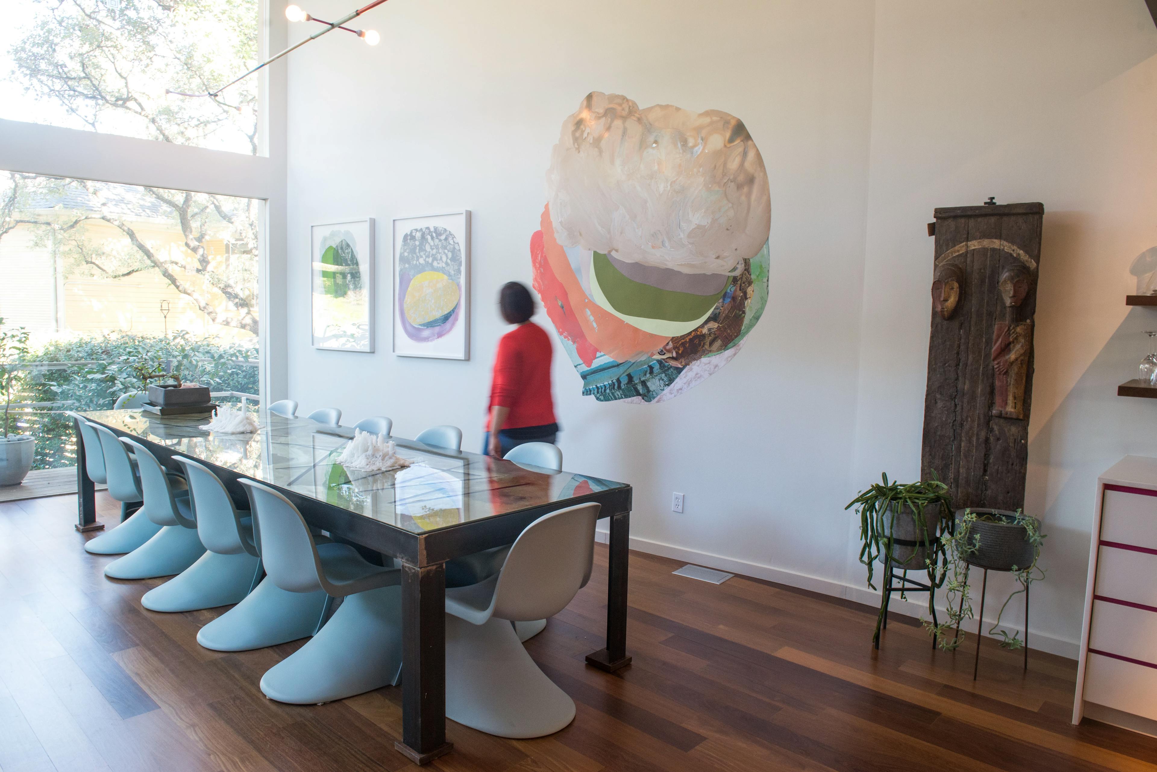 Three original collages by artist Xochi Solis, two framed and one large site-specific work, installed on a white wall in a dining room with a long glass table and blue chairs.