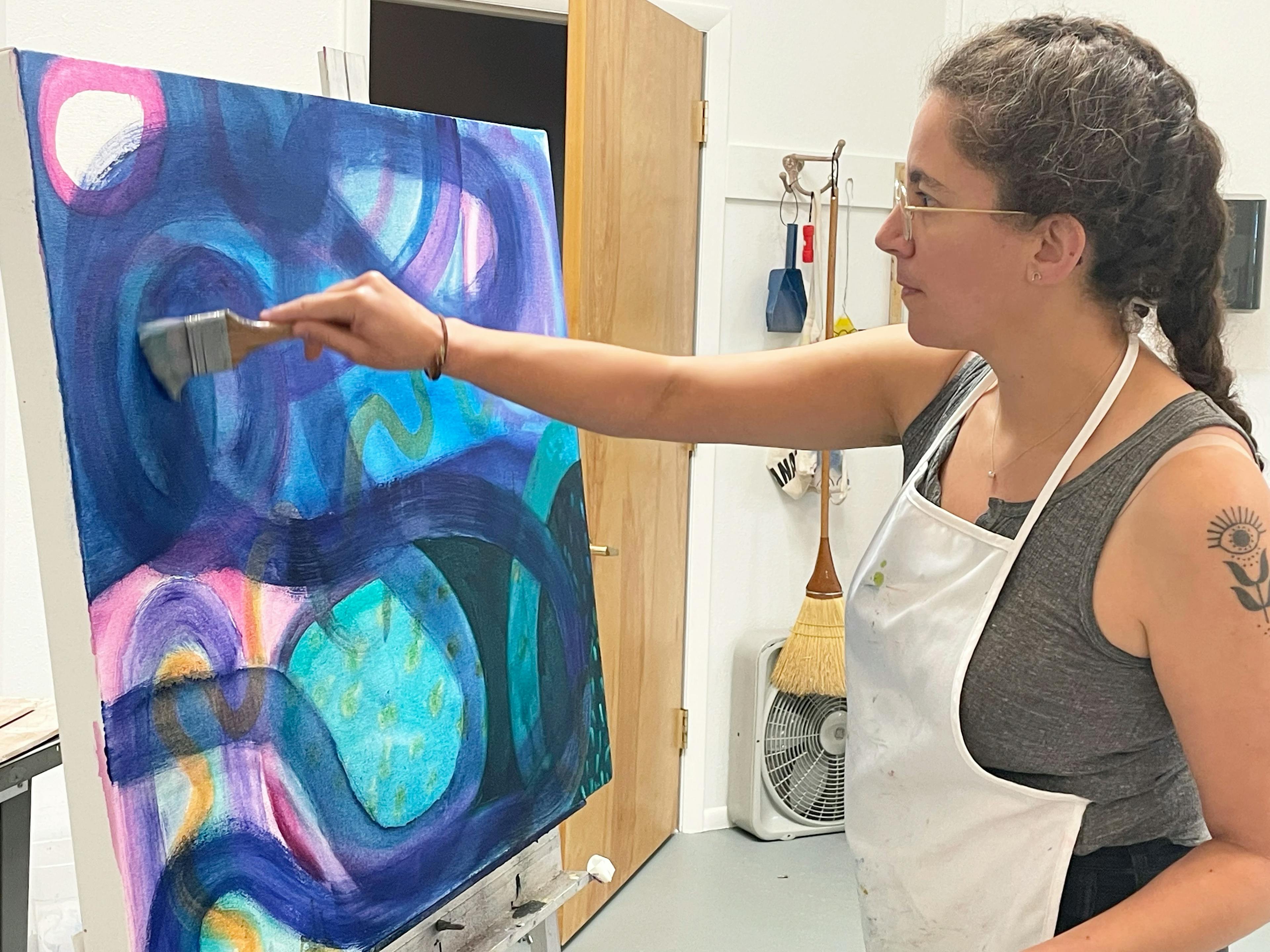 Artist Aliza Cohen wearing an apron and painting dark blue swaths of paint on a canvas in her studio.