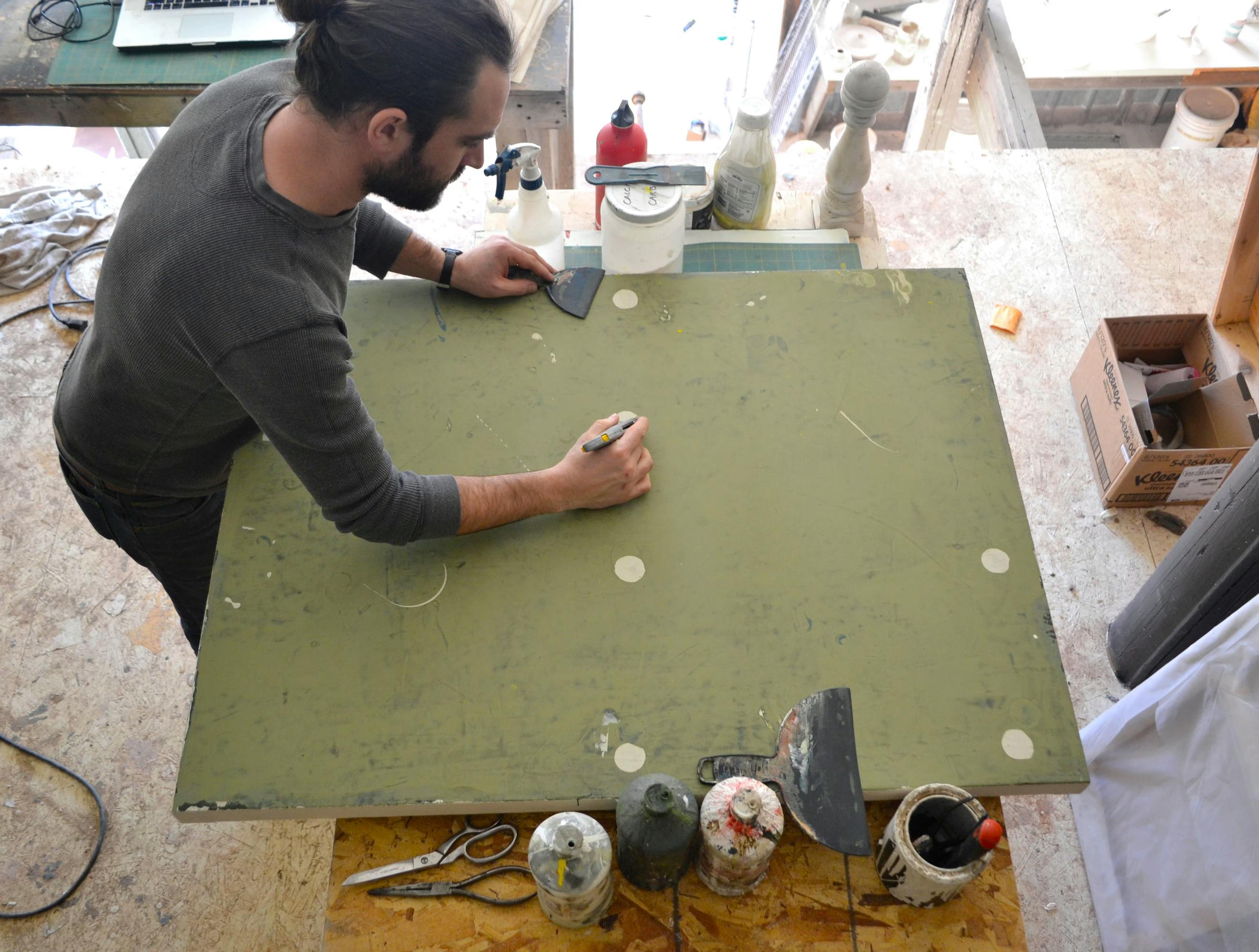 Artist Clay Mahn in his studio working on a distressed green canvas with beige dots.