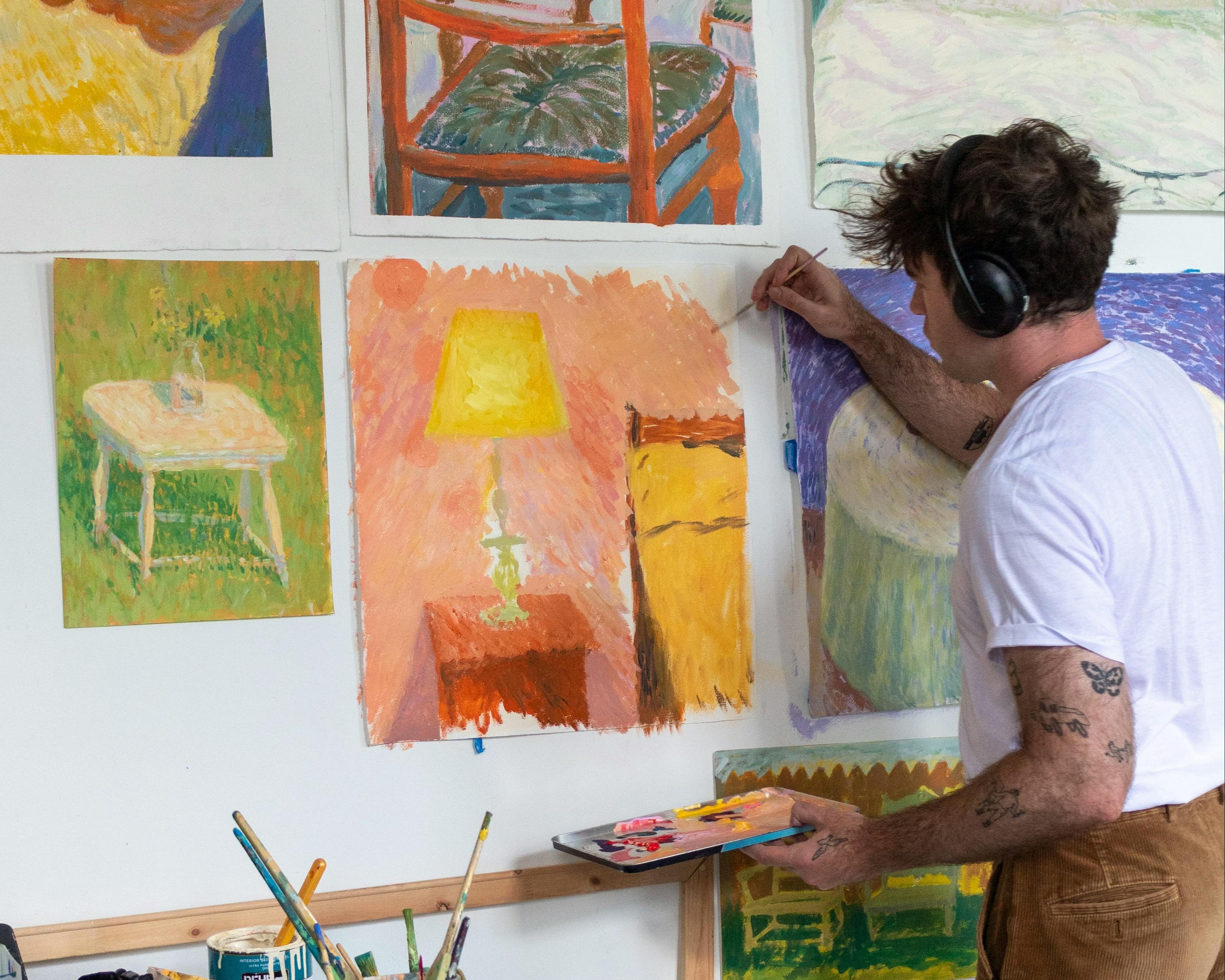 Artist Jackson Joyce wearing over the ear headphones and painting a still life work on paper installed on a white wall in his studio.