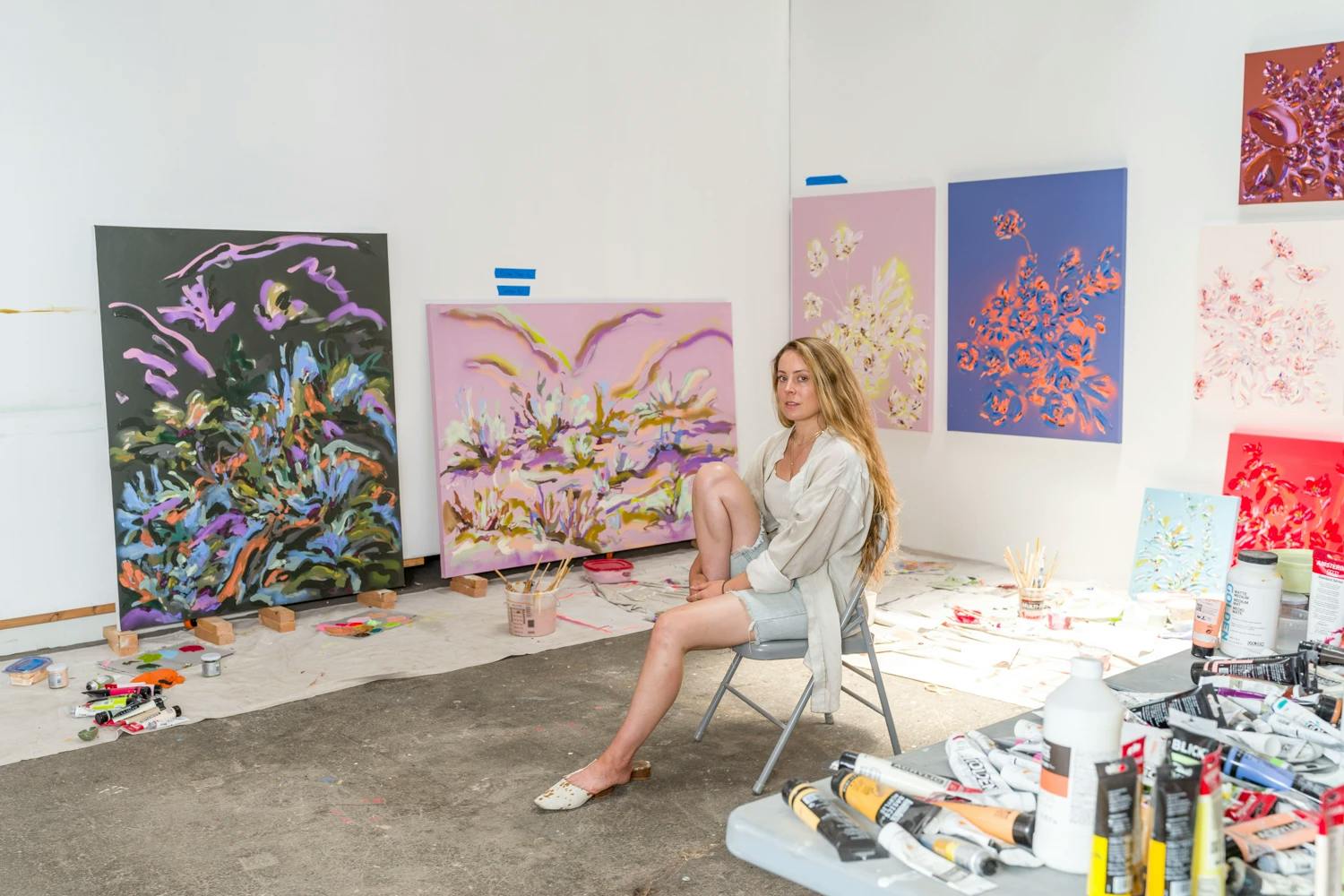 Artist Erin Lynn Welsh sitting on a chair in her studio, surrounded by floral paintings. 