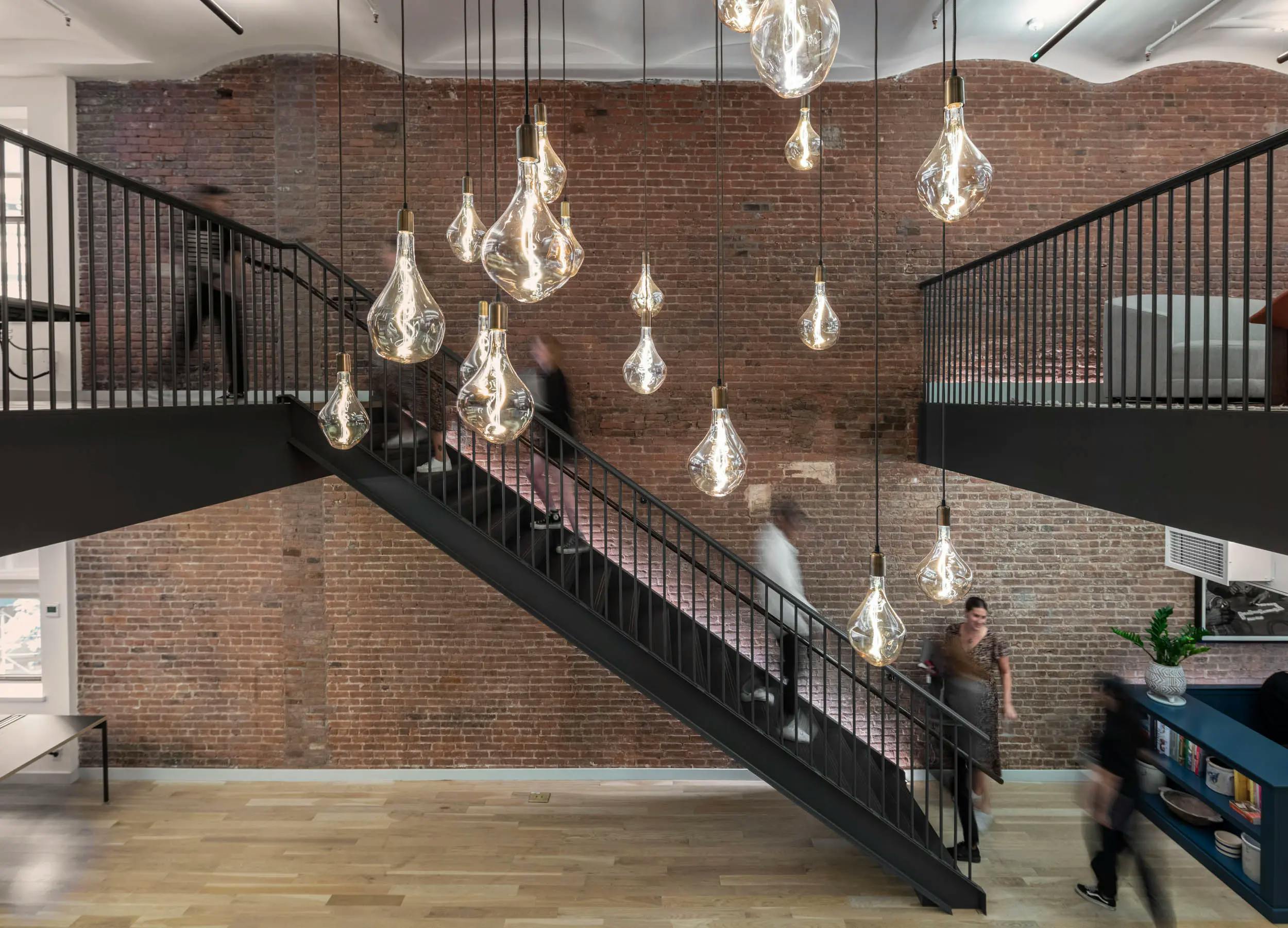 FIG employees walking up an black metal staircase within an open-concept workspace with suspended Edison Light bulbs and a large brick wall.