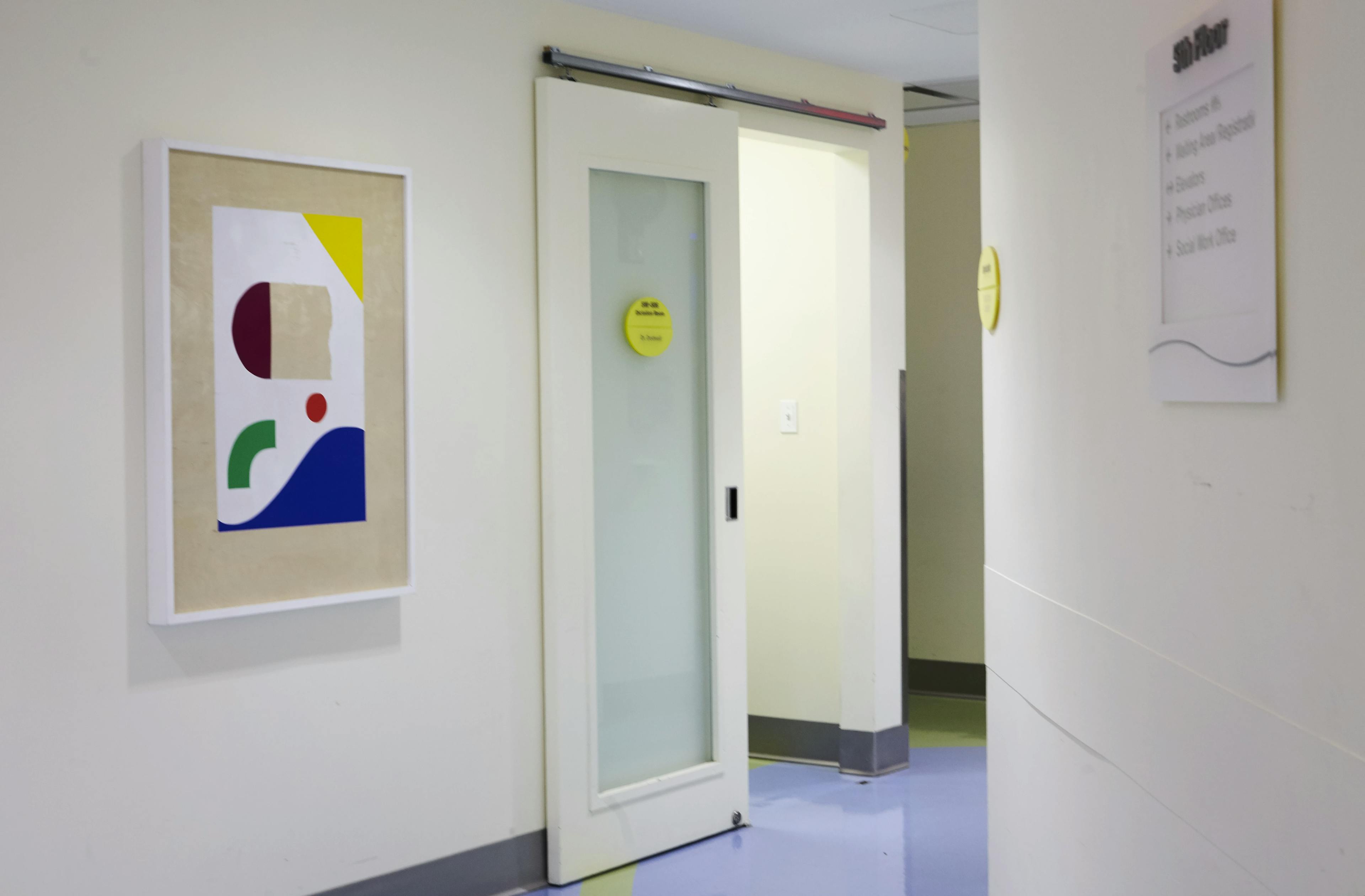 Framed, abstract artwork with geometric shapes installed on a white wall in a hospital corridor.
