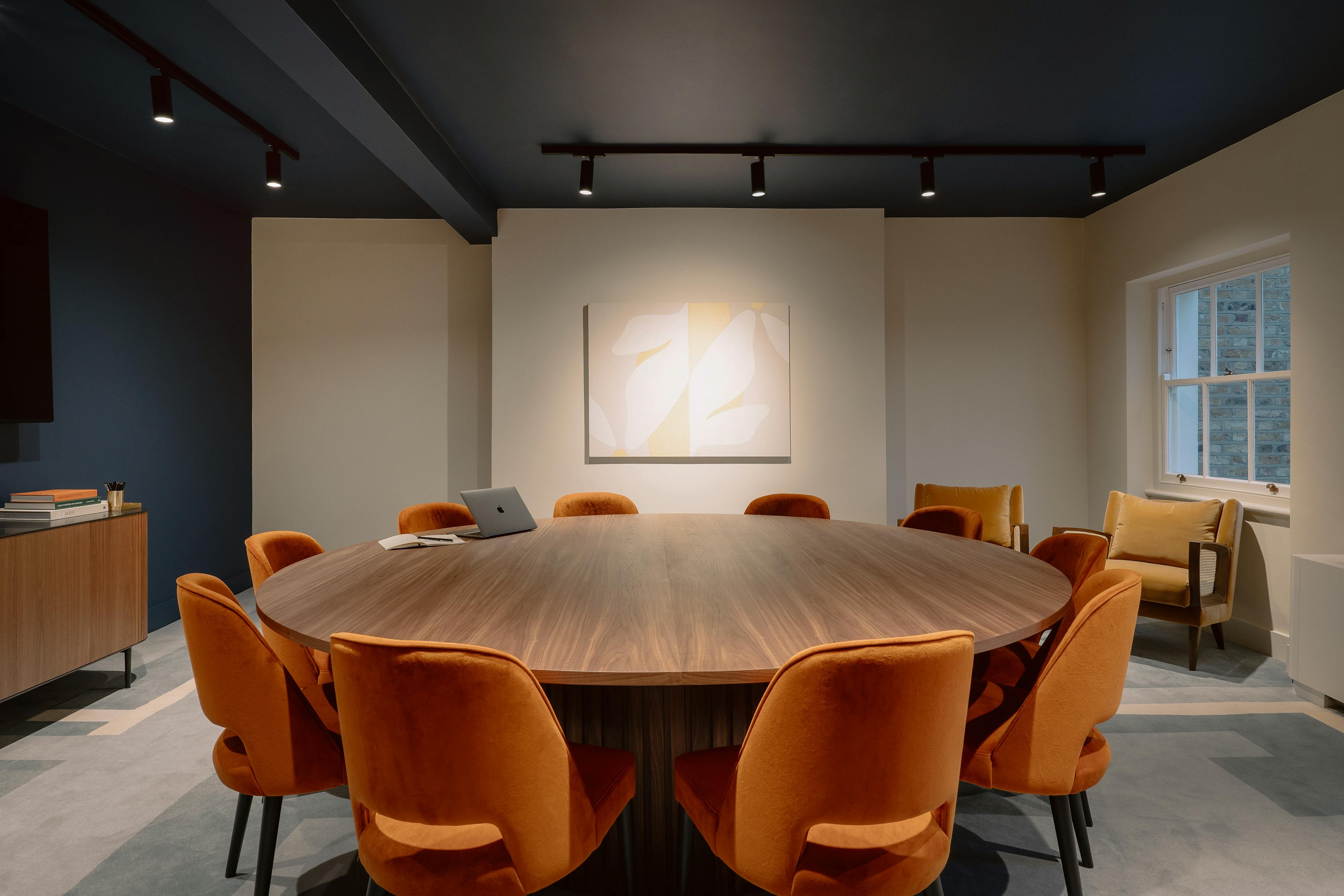 A boardroom at the Chief London clubhouse, featuring a circular wooden table with orange chairs and a neutral floral painting by artist Senem Oezdogan installed on a beige wall.