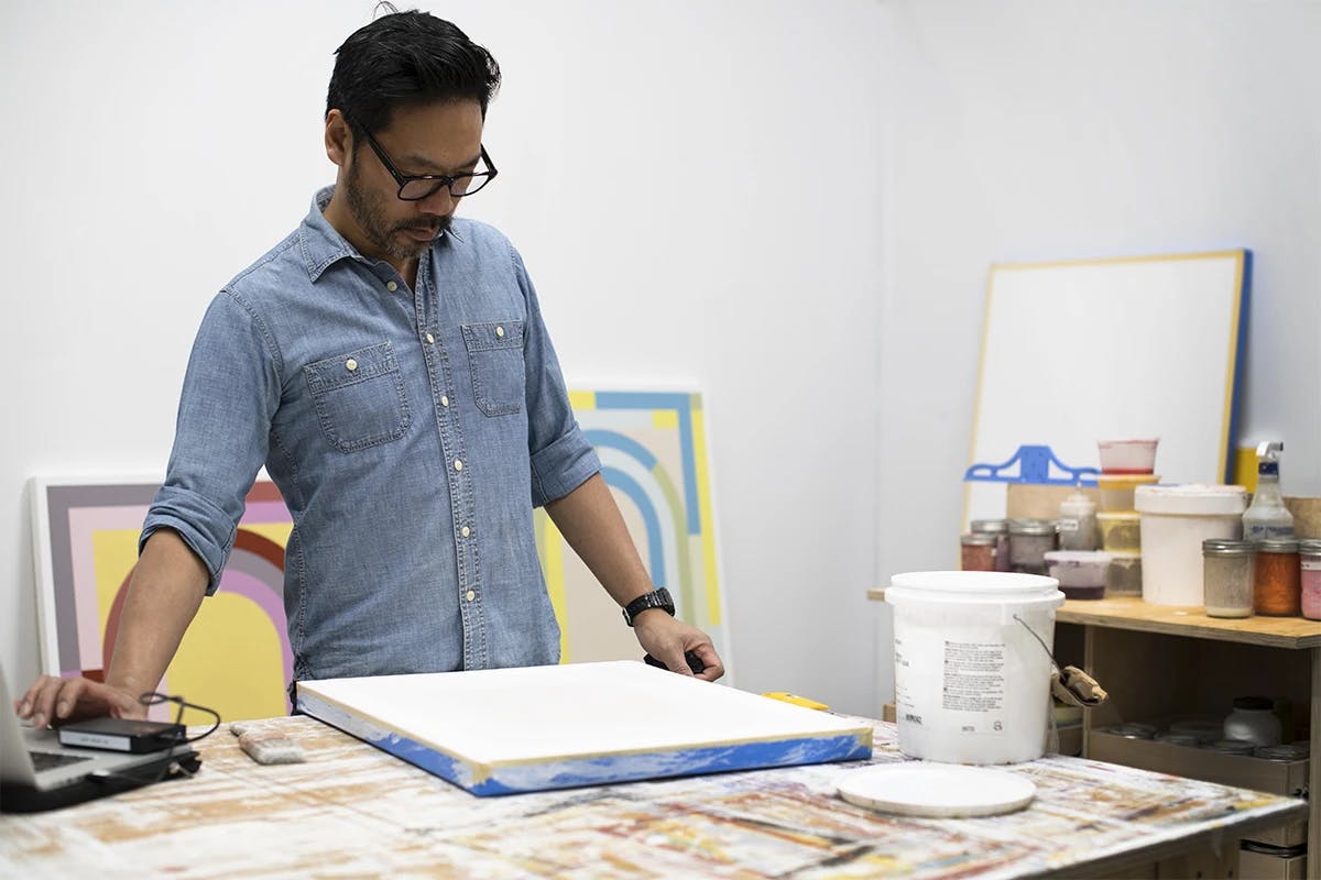 Artist Christian Nguyen wearing a denim shirt, working on his computer while standing in his studio surrounded by paintings on canvas.