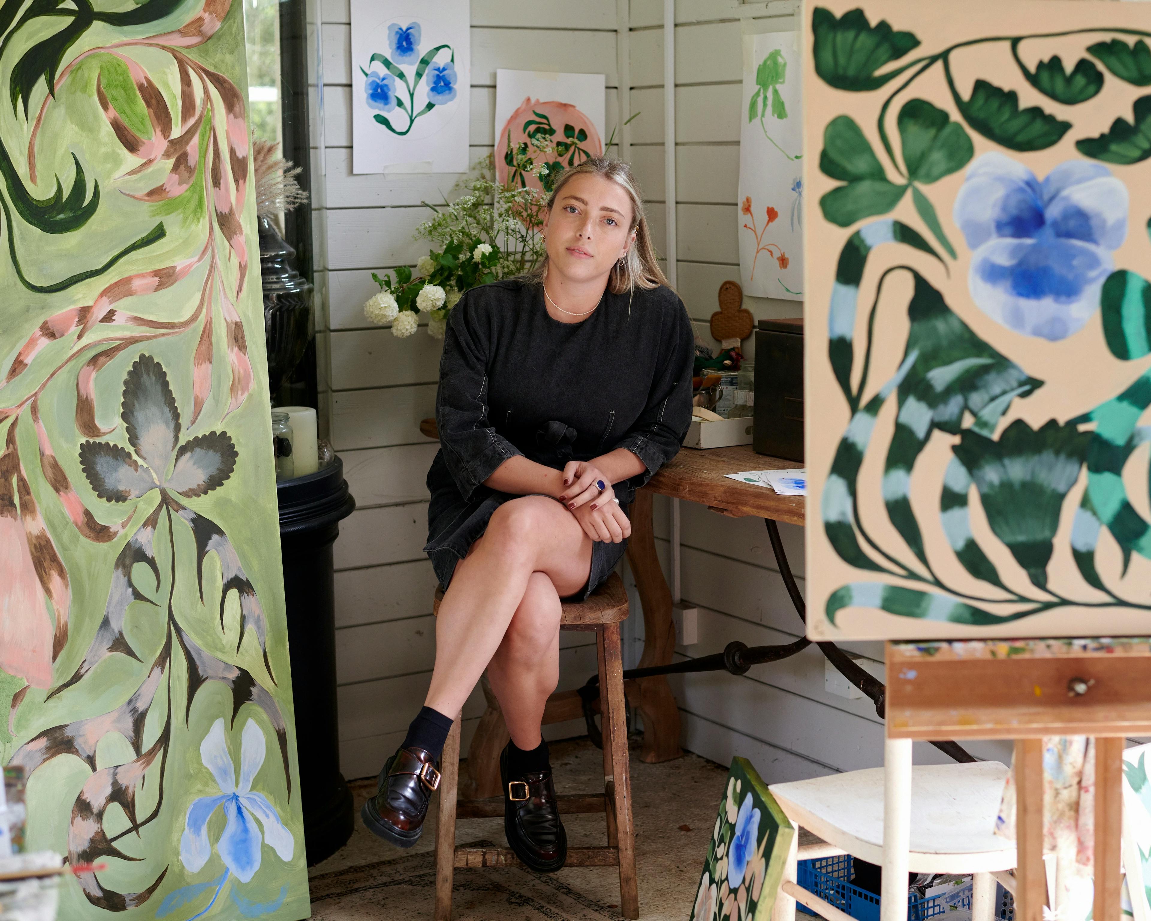 Artist Georgia Beaumont sitting on a stool in her studio, surrounded by her large botanical paintings.