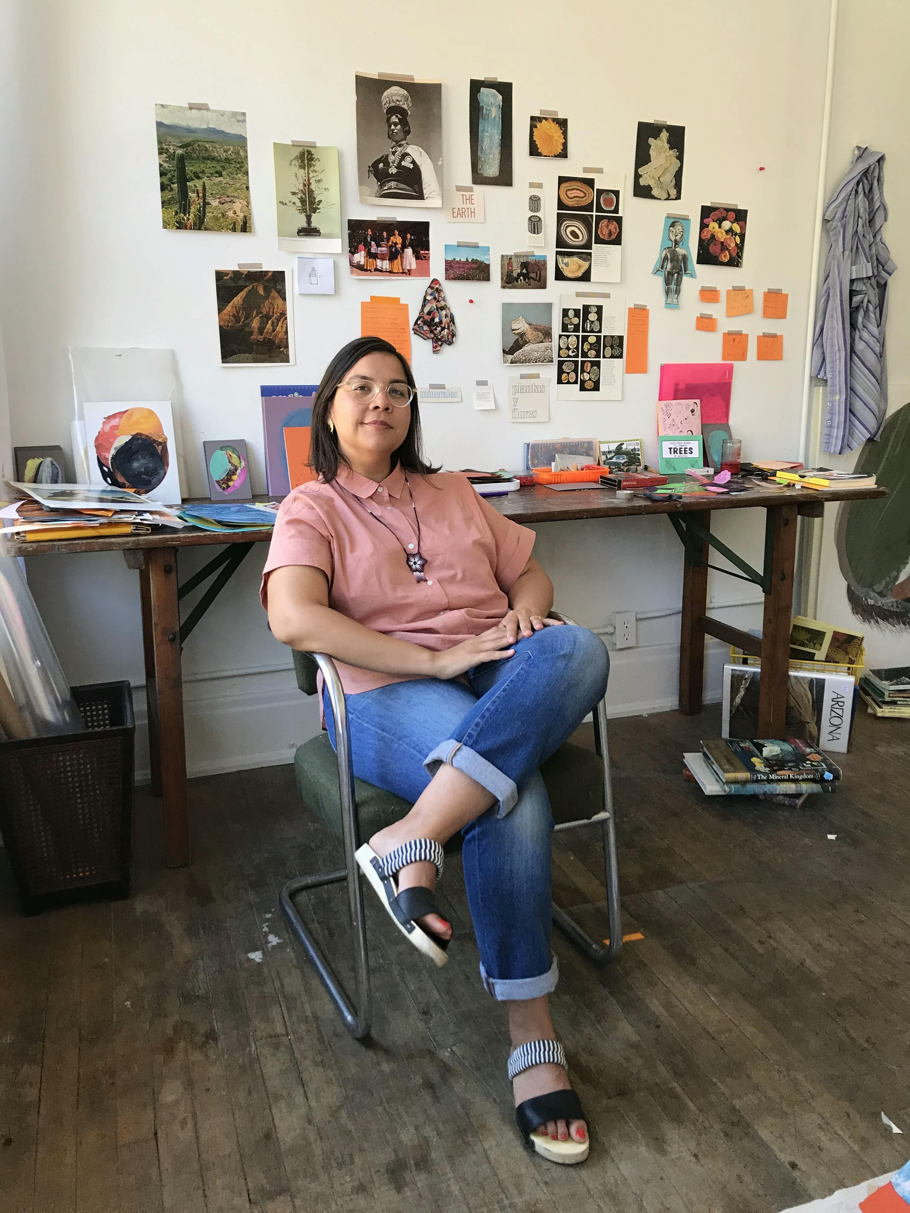 Artist Xochi Solis sitting in her studio surrounded by mixed-media collages and source material in her studio.