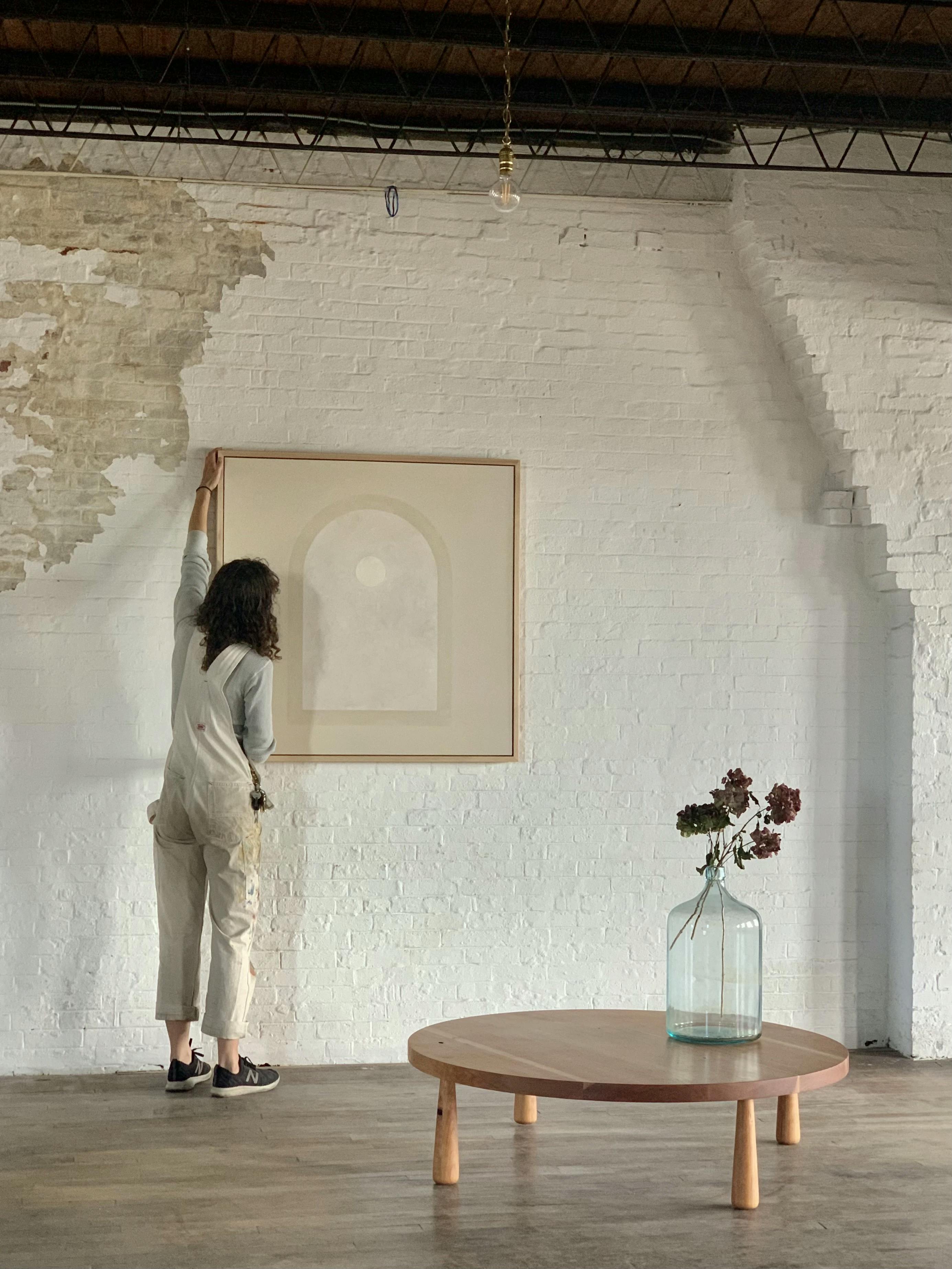 Artist Carla Weeks holding a large, neutral arch painting on a white brick wall.