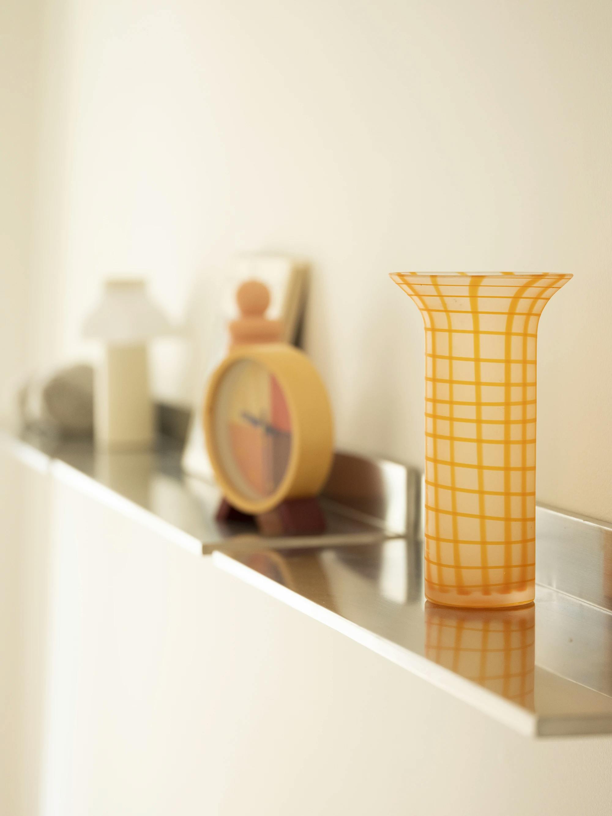 A yellow vase and a clock on a shelf. 