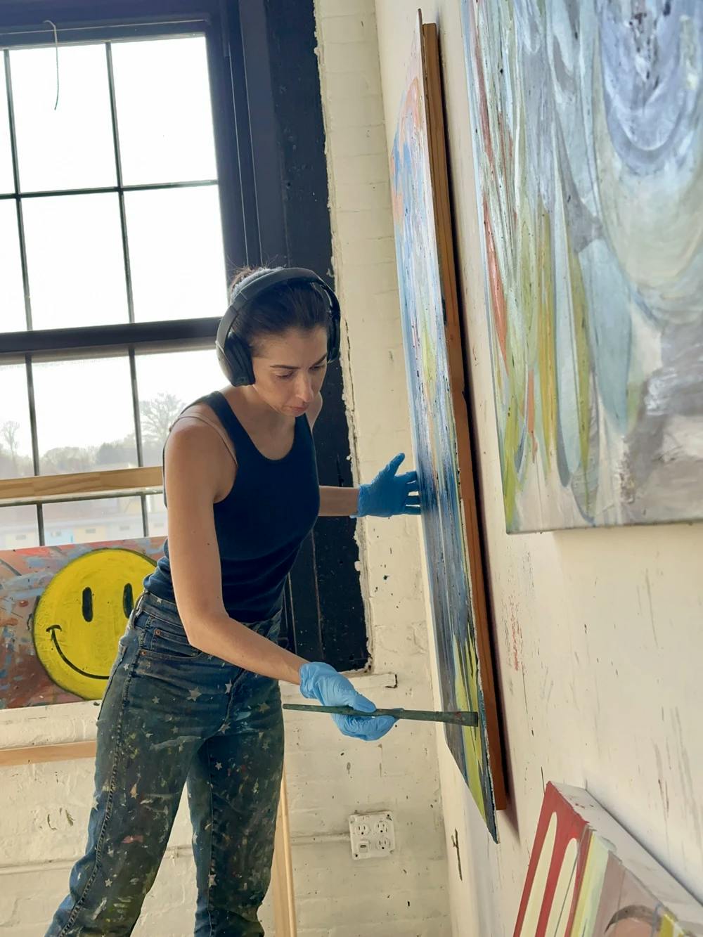 Artist Diana Delgado wearing headphones and wearing plastic blue gloves while painting a large, abstract work.