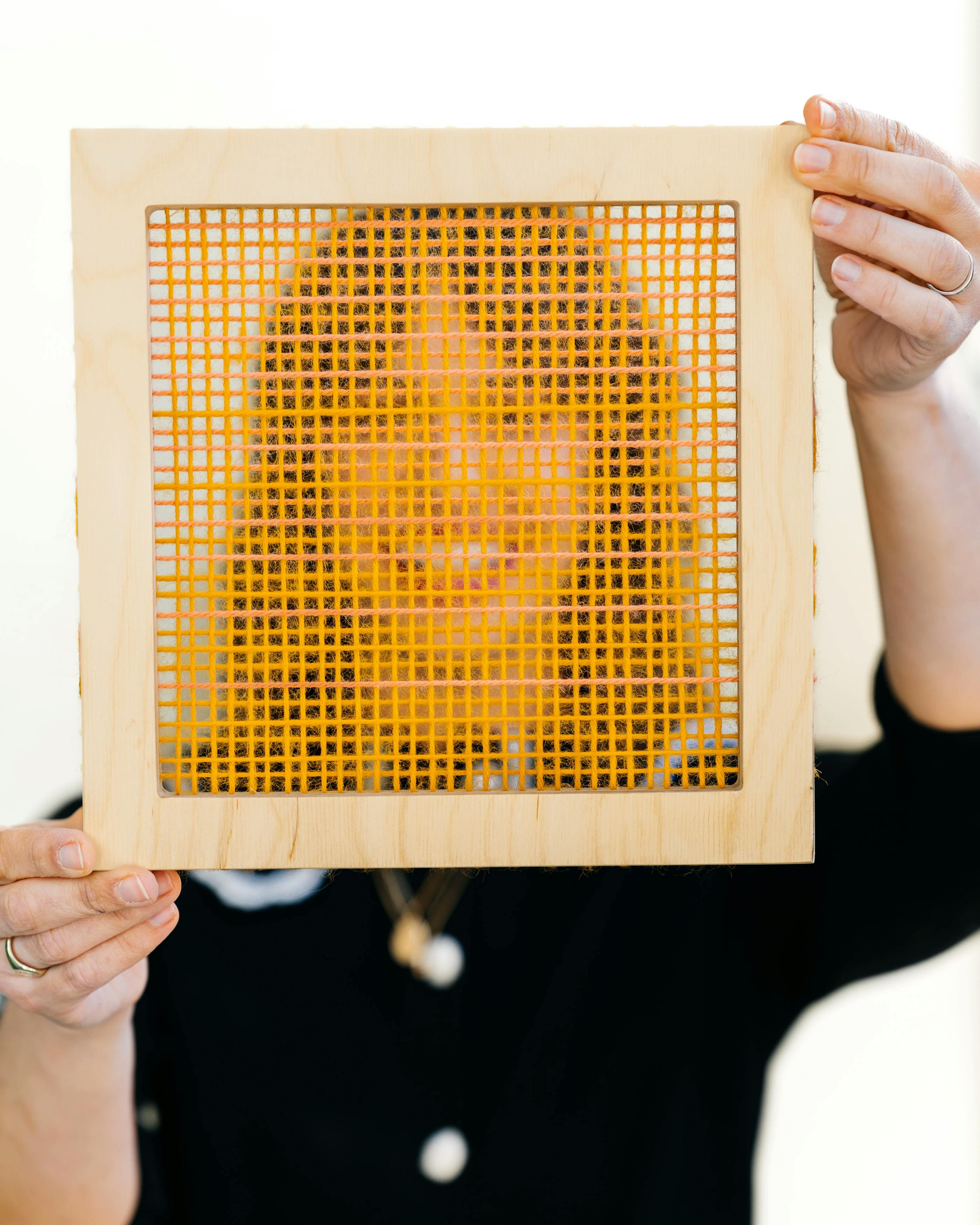 Artist Hayley Sheldon holds a wood frame with yellow thread in front of her face at MacArthur Place.