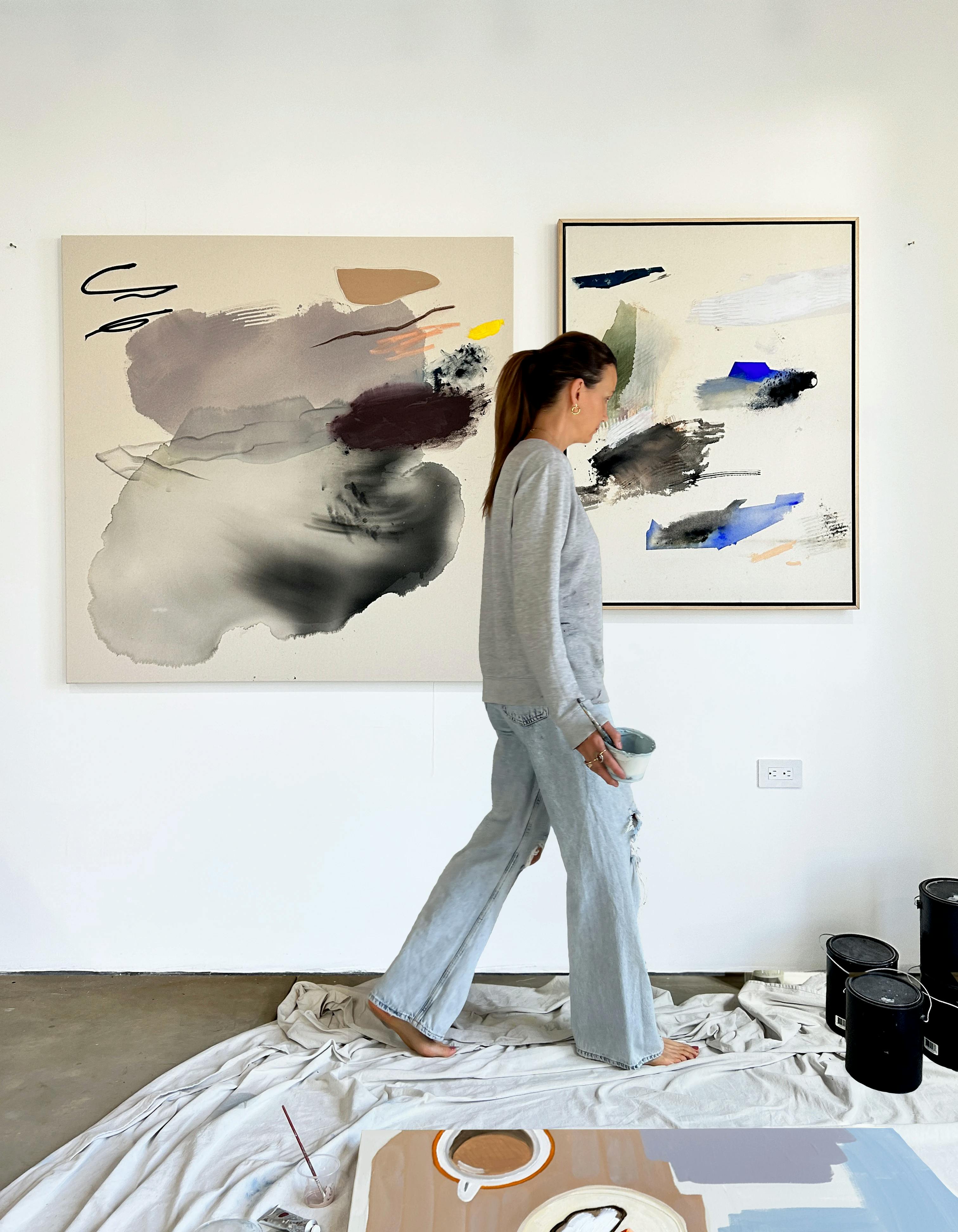 Artist Karina Bania walking in front of two abstract paintings in her studio. 
