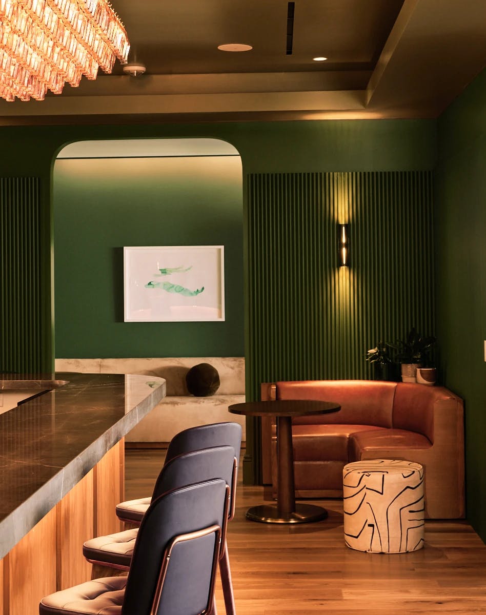 A framed watercolor painting of two figures in repose by artist Gabrielle Raaff installed on a dark green wall above a beige sofa within a bar area at the Chief San Francisco Clubhouse.