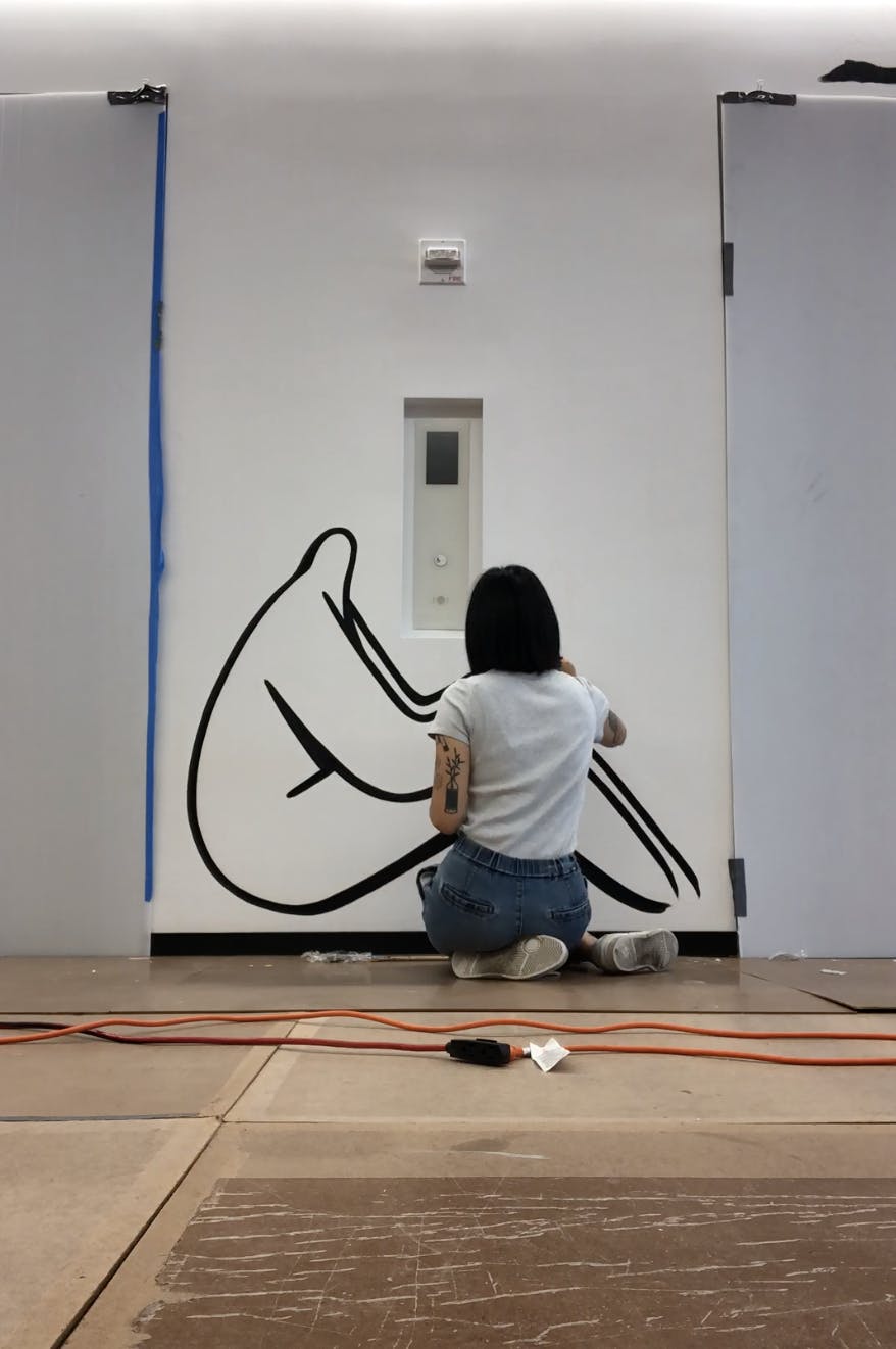 Artist Jocelyn Tsaih sitting on the floor, painting a life-size mural of a black abstract figure.
