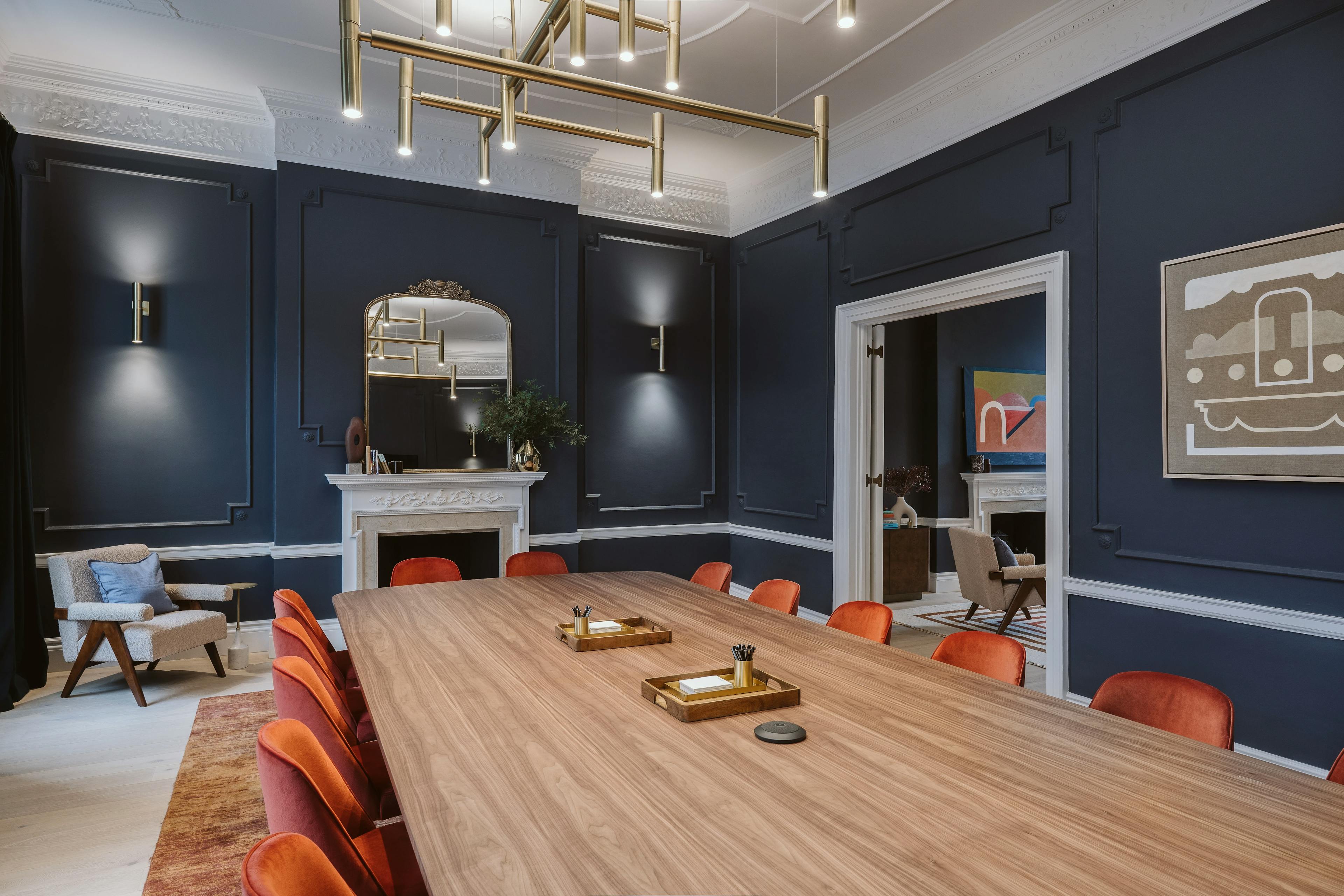 A boardroom at the Chief London clubhouse with a long wooden table, orange chairs, and two paintings by artists Kristin Texeira and Carla Weeks installed on dark blue walls.