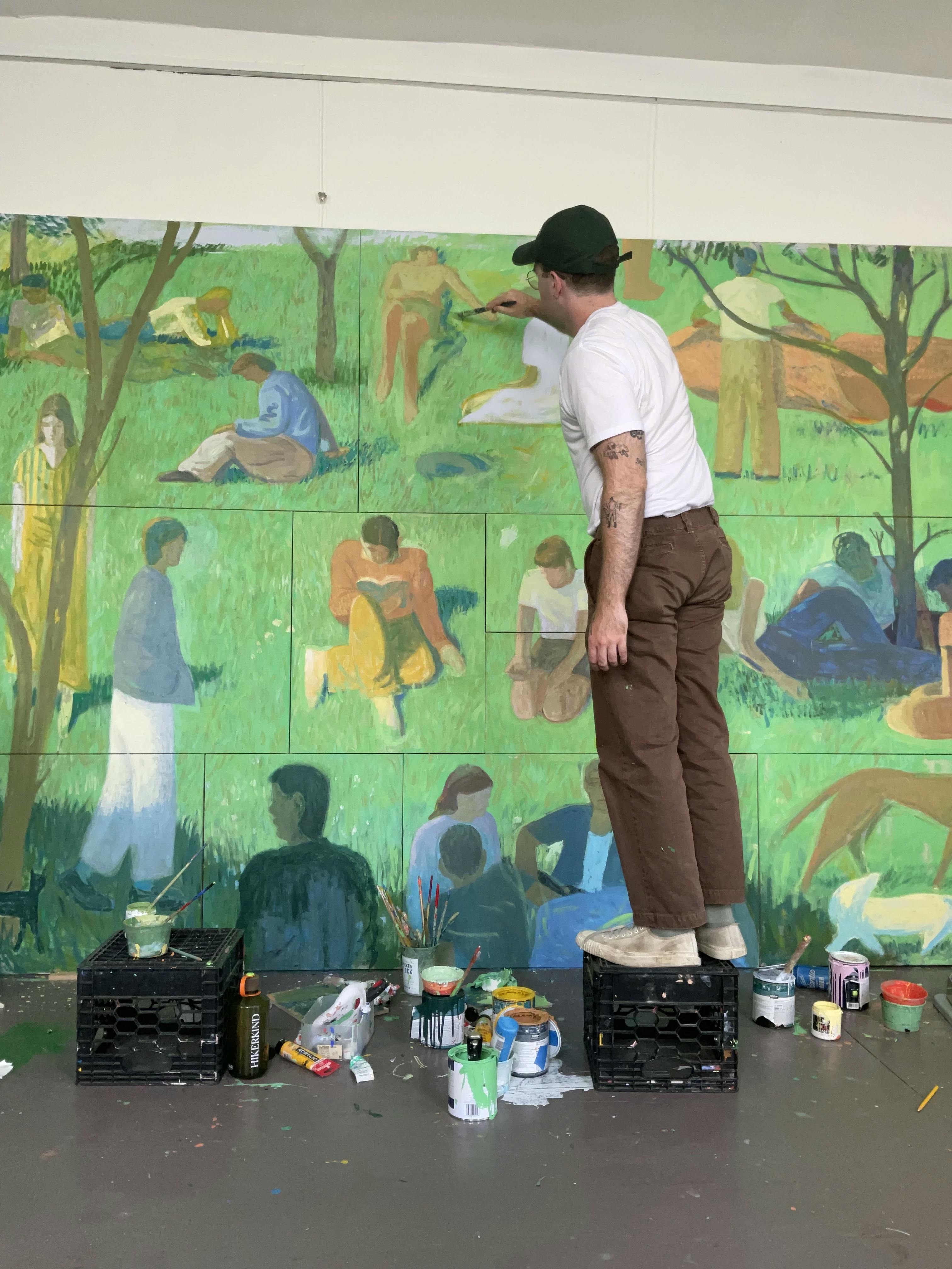 Artist Jackson Joyce in his studio standing on a black box, painting figures on his large-scale installation comprised of individual paintings.