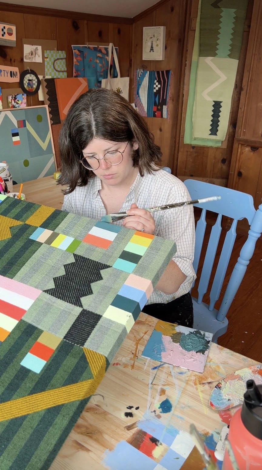 Artist Sarah Sullivan Sherrod sitting a light blue chair, working on a patchwork woven and painted piece in her studio.