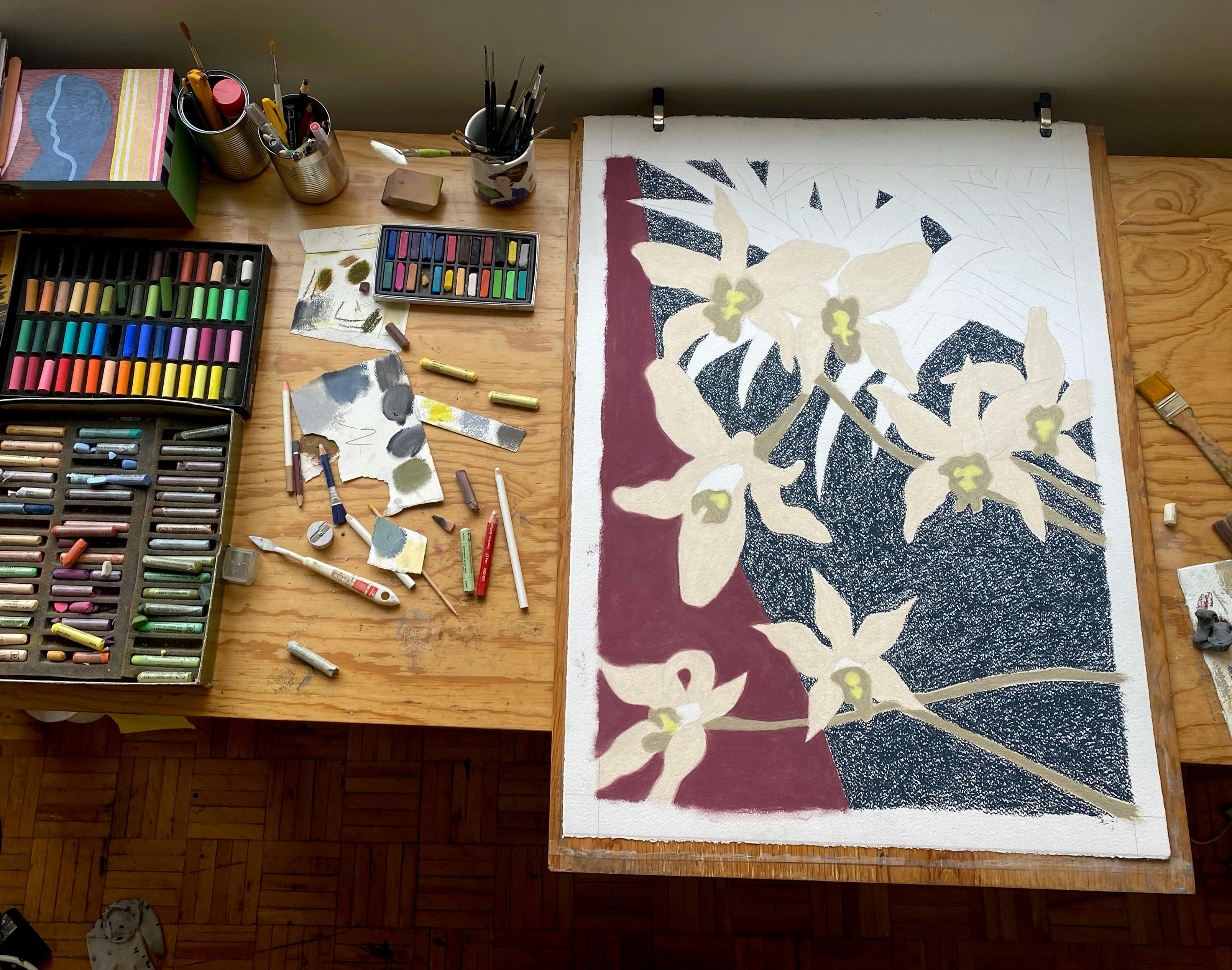 A wooden table in artist Rachel Levit Ruiz studio, covered with boxes of pastels and an in-progress drawing of a flower.