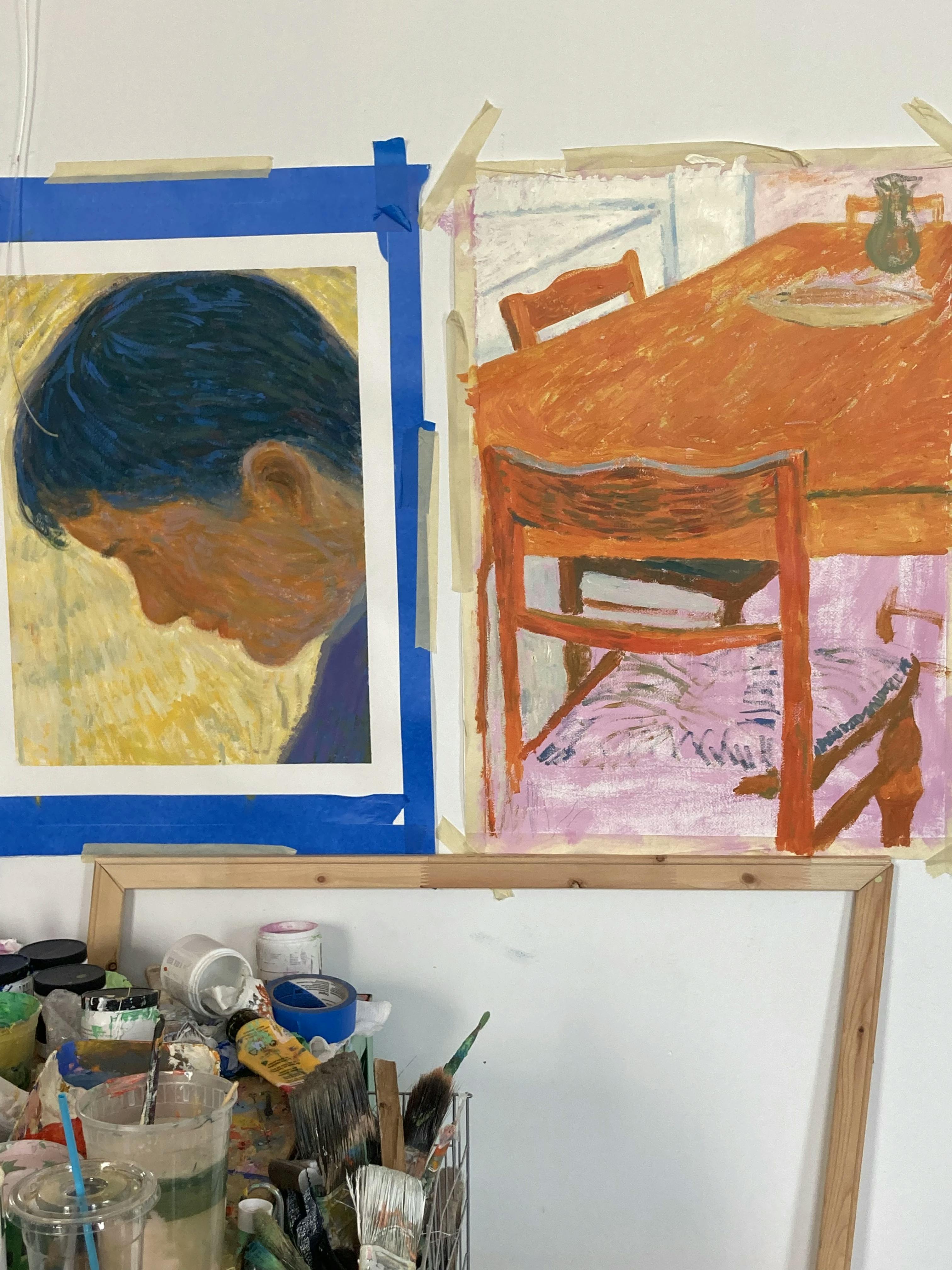 Two paintings by artist Jackson Joyce, one a portrait of a man and the other of a wooden table, taped to a white wall in his studio.