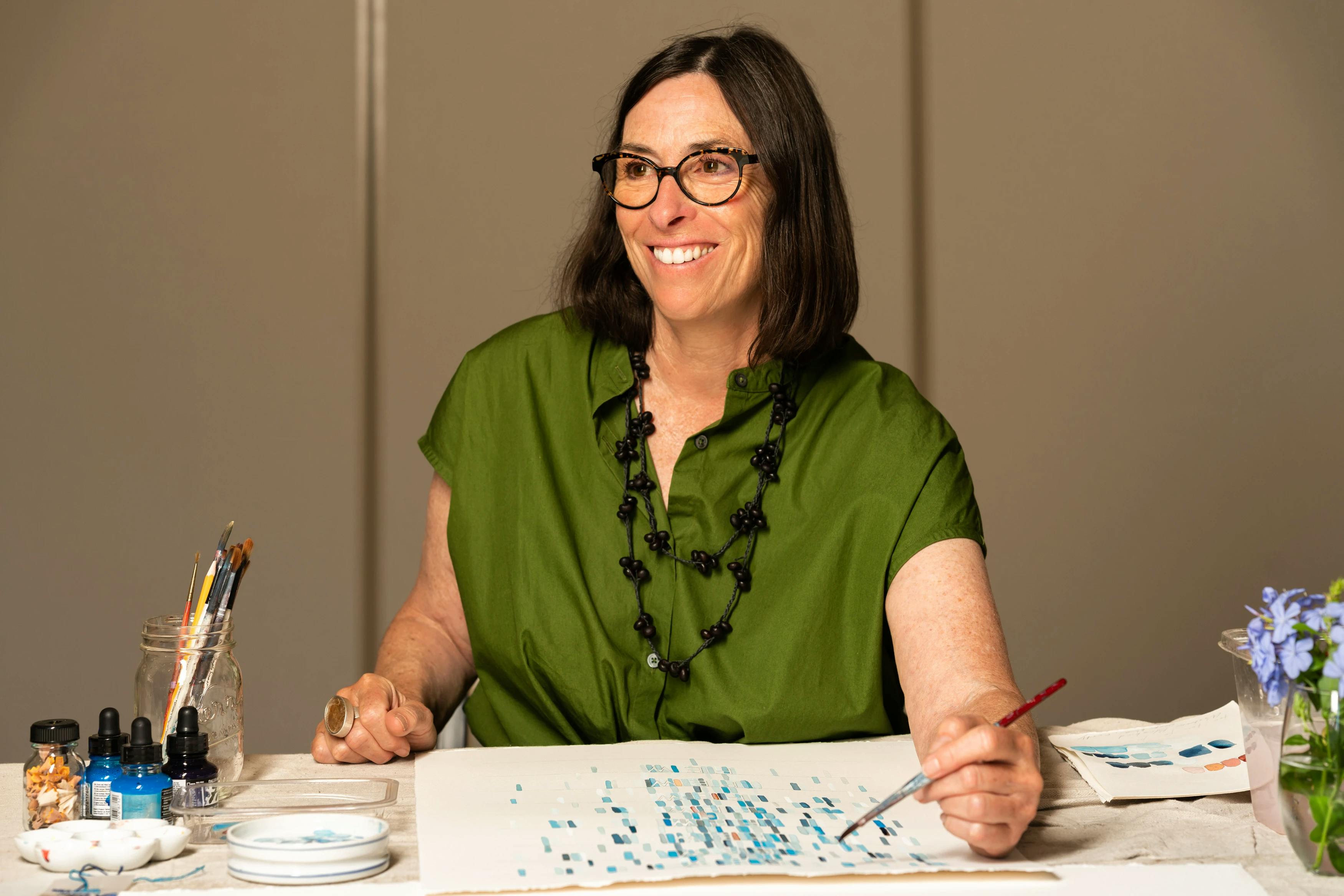 Artist Gail Tarantino wearing a green shirt and holding a paintbrush within a studio at MacArthur Place.
