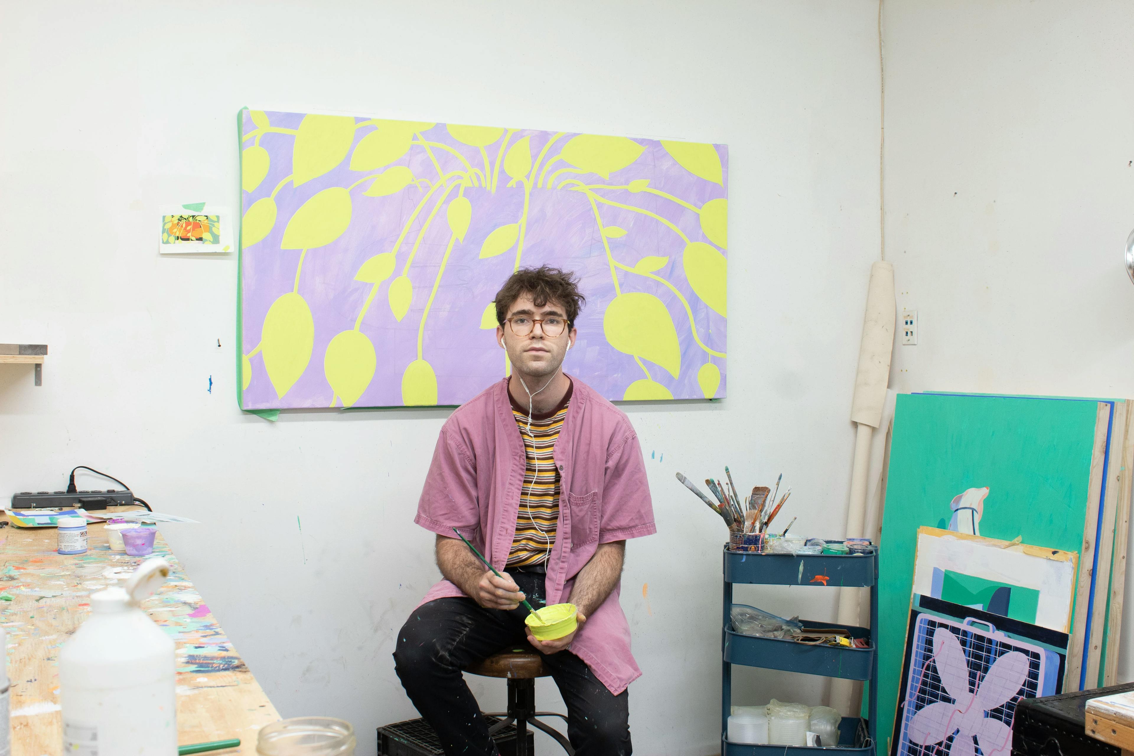Artist Jackson Joyce sitting on a stool in his studio surrounded by his colorful paintings.
