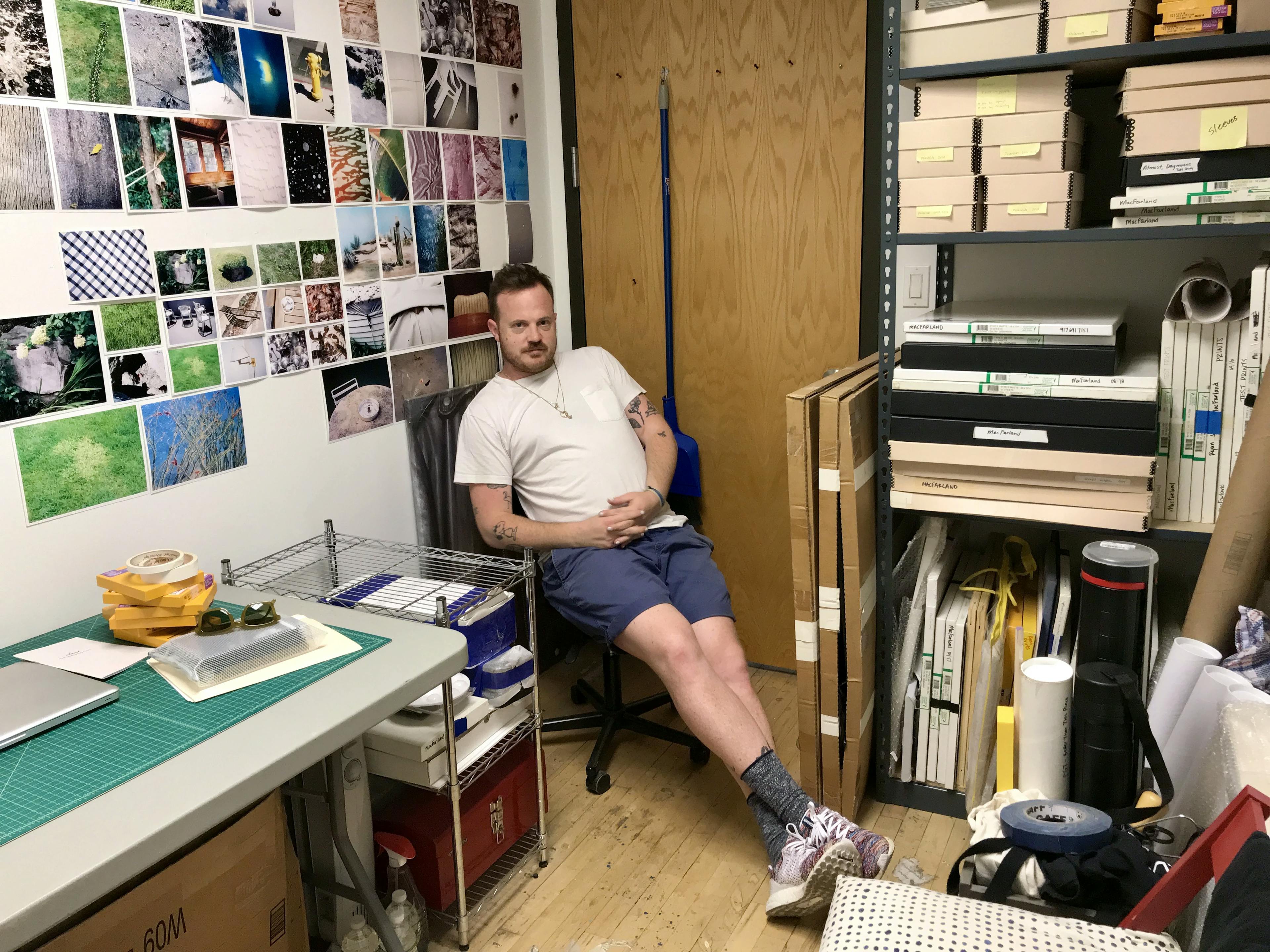 Artist Ryan James MacFarland sitting on a chair in his studio surrounded by his photographs pinned up in a grid on a white wall.